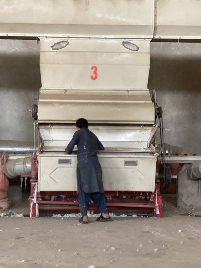 A man stands facing a large, off-white industrial machine with the number "3" in red on its upper section. The man is wearing a dark blue tunic and loose pants, with dark sandals on his feet. He appears to be working on or inspecting the machine. The machine is part of what seems to be a textile or cotton processing facility, judging by the scattered tufts of cotton on the floor and around the base of the machine. The setting appears to be indoors, with pipes and parts of other machines visible in the background. There's nothing in the image to indicate it's specifically in Kandahar, Afghanistan.