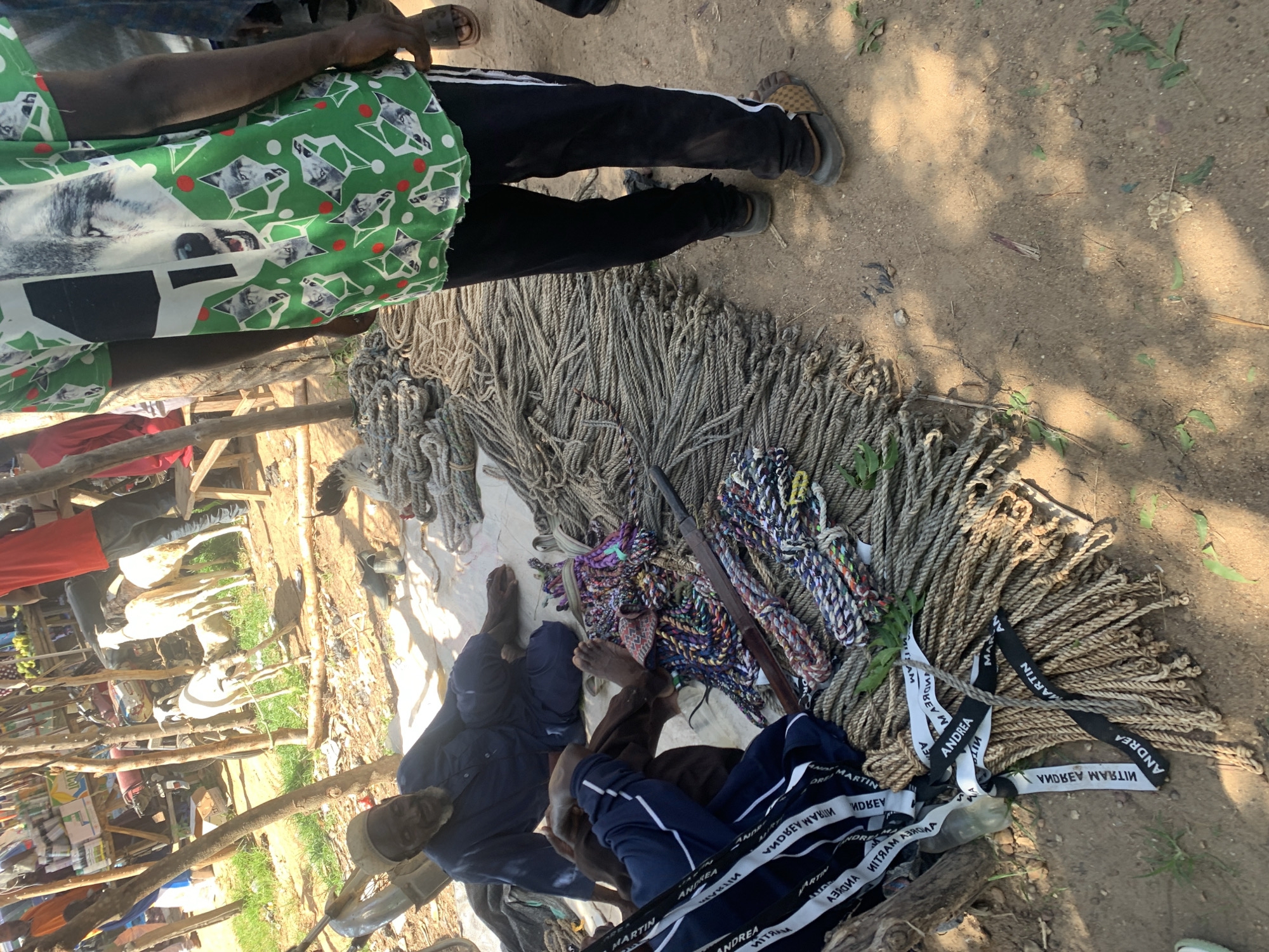 This image captures a scene at what appears to be an outdoor market or gathering, possibly in Nigeria, given the project context. The foreground is dominated by a large pile of coiled ropes of various sizes and colors. Some are thick and natural-toned, while others are thinner and brightly colored in patterns of blue, purple, and white.
Interspersed with the ropes are black and white fabric strips with text printed on them. The visible text includes words like "ANDREA," "MARTIN," and "MITRAM," repeated multiple times, some in reverse.
Several people are present. In the upper part of the image, a person is seen wearing a green t-shirt with a wolf print and black pants with white stripes. Their legs and feet, clad in sandals, are visible. Further down, two individuals are seated amongst the ropes, their legs bent. One is wearing dark blue pants with white stripes, and the other is in dark blue pants and a dark shirt. Their faces are partially visible, but their expressions are not distinct.
In the background, a wooden structure is visible, suggesting stalls or shelters typical of a market. Goats and possibly other livestock can be seen behind this structure, indicating a rural or semi-rural setting. The ground is dusty and sandy, with patches of dry leaves and sparse vegetation. The lighting suggests it is daytime, with shadows indicating the sun's position. The overall atmosphere seems relaxed, with people possibly resting or attending to the merchandise.