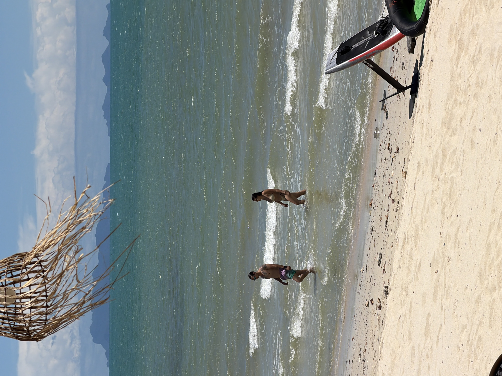 Two individuals, a man and a woman, walk through shallow water on a sandy beach in Baan Tai, Thailand. The man, wearing colorful swim trunks, is on the left, and the woman, in a bikini, is to his right and slightly behind. The ocean water is a greenish-blue with small waves. In the foreground, the sandy beach is scattered with small rocks and debris. To the right, a hydrofoil surfboard is propped up on the sand, partially obscuring a black inflatable object. In the background, the sea extends to the horizon, with a line of mountainous islands visible under a partly cloudy sky. A wicker lampshade hangs in the upper left corner of the frame, obscuring part of the sky and the distant islands.