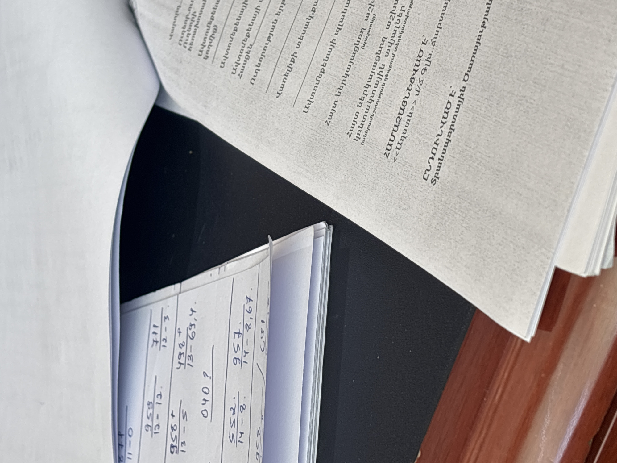 The image shows a close-up view of papers with handwritten mathematical calculations and text in Armenian. One paper has columns of numbers with operations like addition and subtraction, and some numbers are underlined. Another paper, opened to the right, has printed Armenian text, with some lines being longer than others, suggesting it's a document or book page. The papers are placed on a dark surface, possibly a desk, with a wooden surface visible at the bottom left. The lighting suggests an indoor environment. There is no indication of a specific location in Ijevan, Armenia.