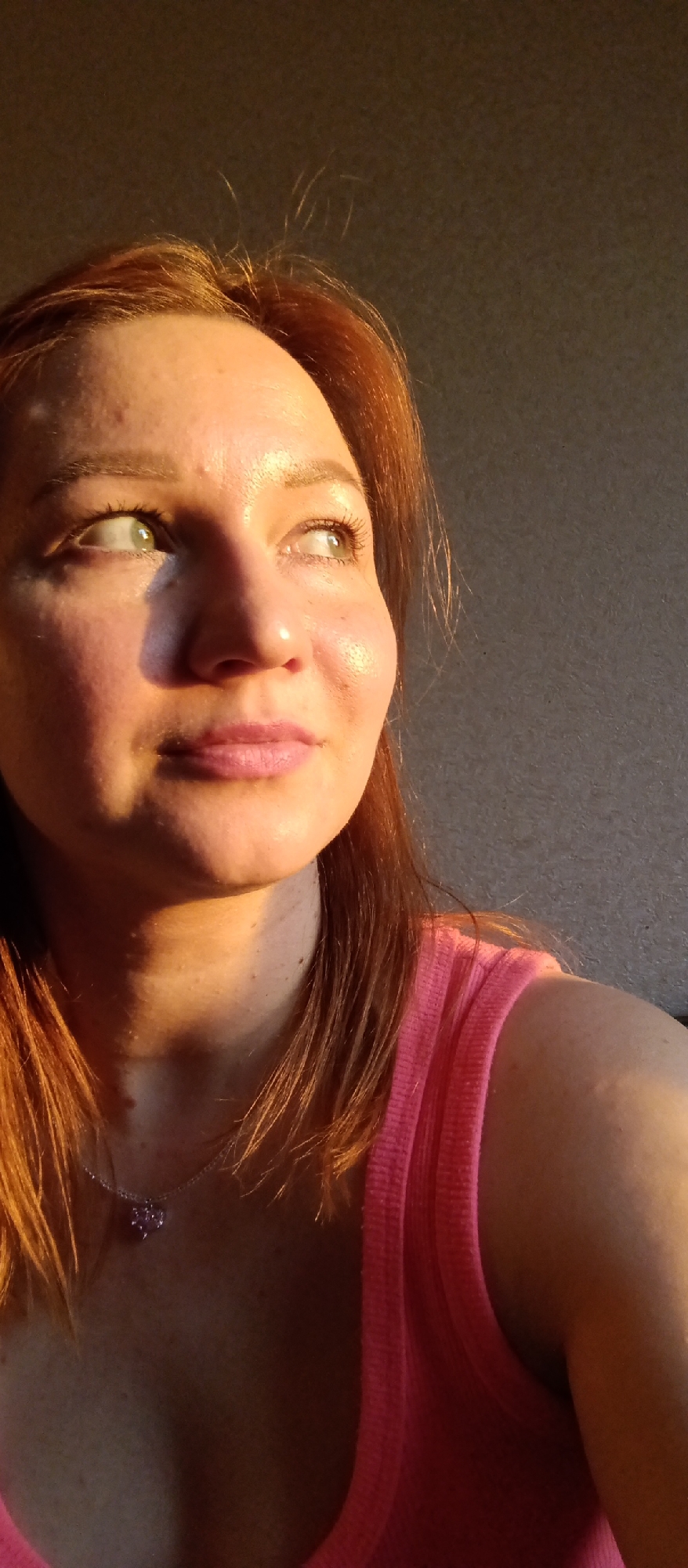 A close-up shot depicts a Caucasian woman with reddish-brown hair, light skin, and green eyes. She is wearing a pink tank top and a necklace with a pink pendant. The woman is looking upwards and to the left, away from the camera. The lighting is warm and golden, suggesting it might be late afternoon or early evening. The background is a neutral grey wall. The location is not specified in the image.