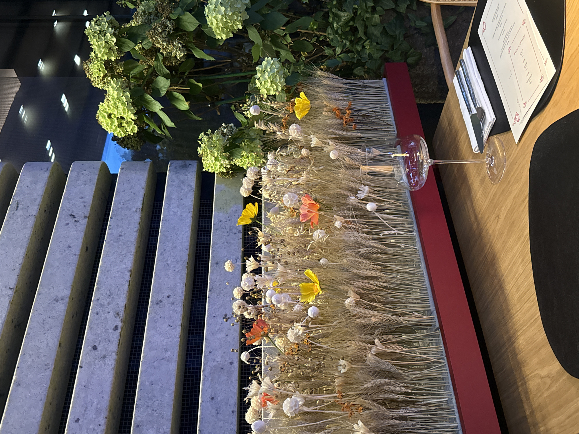 A wide shot captures a table setting in an indoor environment, possibly a restaurant, with a focus on a floral and wheat arrangement. The arrangement, housed in a red planter box, features dried wheat stalks interspersed with delicate white dried flowers, and a few scattered artificial poppies in orange and yellow. Behind the arrangement, a modern architectural element composed of stacked, light-colored stone slabs with horizontal openings is visible. To the right of the stone structure, a cluster of vibrant green hydrangeas and darker foliage adds a touch of natural color. The foreground of the image is dominated by a wooden table. On the table, a single clear wine glass with a long stem stands to the left, and to its right, a white napkin is folded with a knife and fork placed on top. A menu, with red decorative borders, is laid out on a black placemat. The lighting suggests an evening or dimly lit setting, with reflections of ceiling lights visible in the background. No people are present in the scene.
