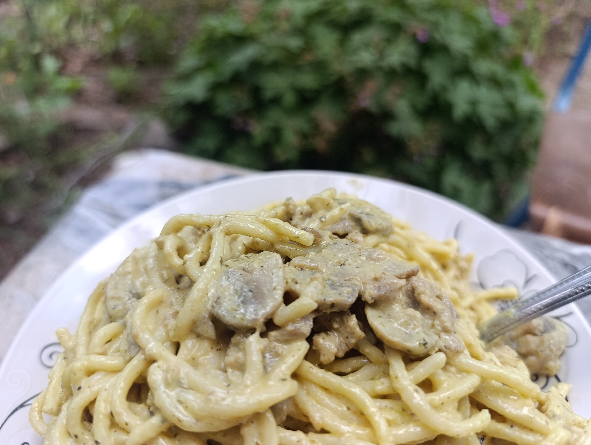 A close-up, high-angle photograph captures a serving of pasta on a white plate. The pasta consists of spaghetti-like noodles thoroughly coated in a thick, light beige creamy sauce containing dark specks, indicative of ground pepper or dried herbs. Mixed within the pasta and sauce are numerous slices of mushrooms and smaller, irregularly shaped pieces of cooked meat. The white ceramic plate features a decorative black swirl pattern partially visible on its left side. A silver-colored metal fork is partially submerged into the pasta on the right side of the plate. The background is heavily blurred, depicting dense green foliage consistent with an outdoor or garden setting, with faint hints of pink or purple flora discernible within the blurred greenery. An indistinct blue object is minimally visible on the far right edge of the blurred background, possibly part of an outdoor structure. The scene is illuminated by soft, natural light, creating a shallow depth of field that focuses sharply on the food. The image provides no visual indicators to confirm the specific location within the city of Nikolichevtsi, Bulgaria.