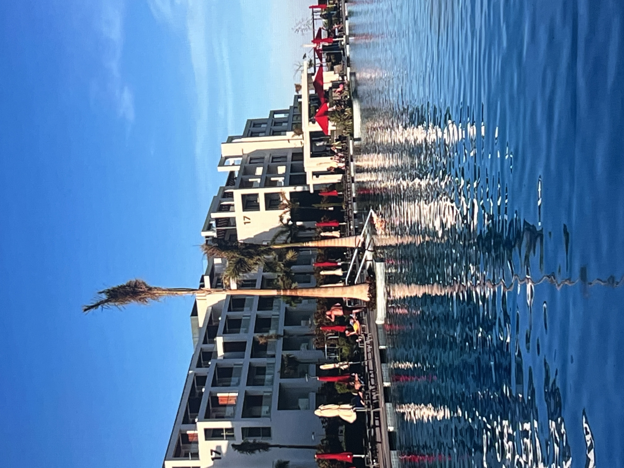 This is a photograph of a resort or hotel with a swimming pool in the foreground. The building is modern, with white facades and multiple balconies. There are several palm trees visible, some of which appear to be dead or dried out. Red umbrellas and lounge chairs are set up around the pool area. Several people are seen relaxing on the chairs and some are standing. The sky is clear and blue. The image is taken from a low angle, looking across the pool towards the building. There is no indication that this location is in Vanadzor, Armenia.