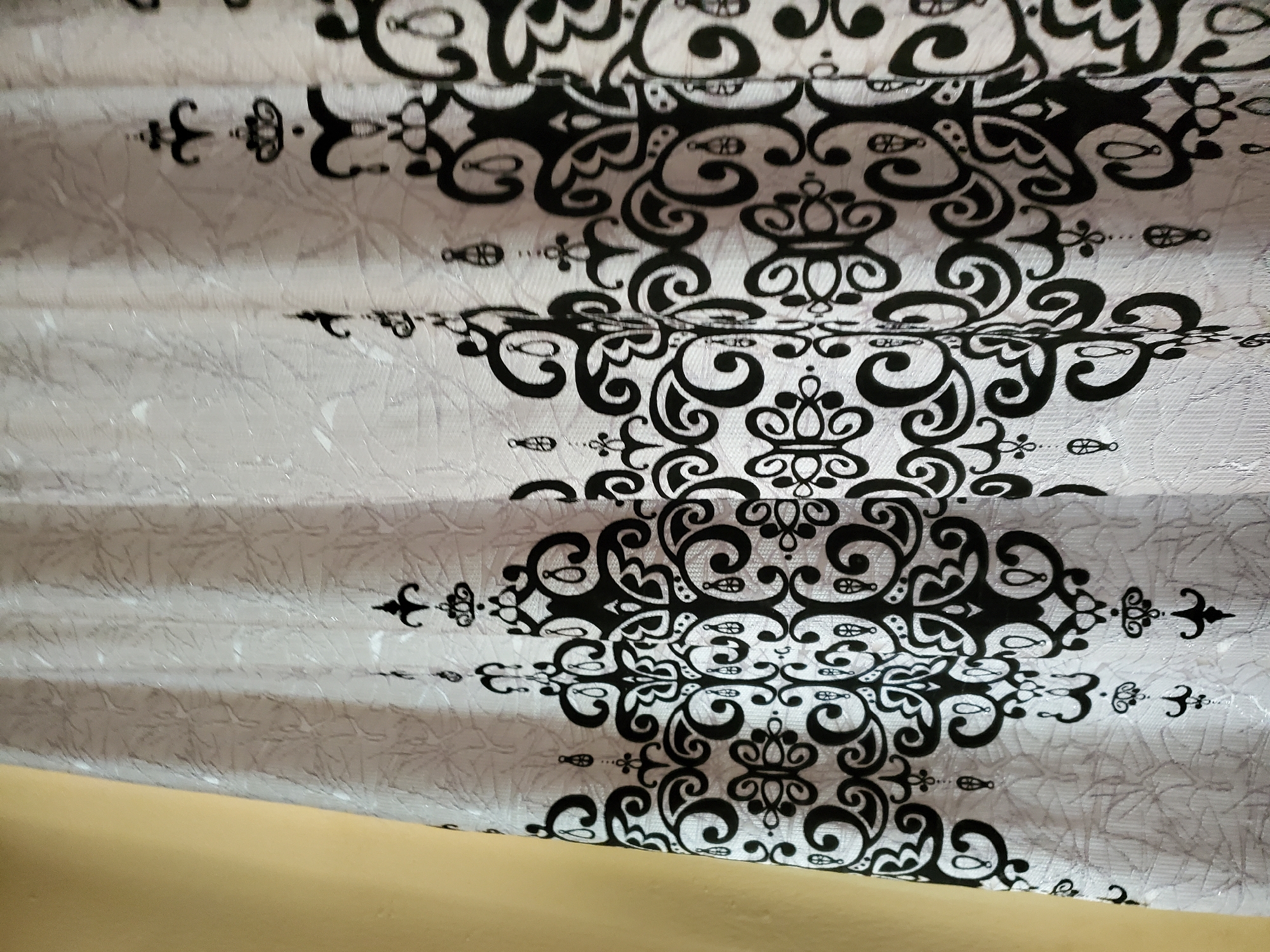 The image shows a close-up of a white curtain with a repeating black ornate pattern. The curtain is hanging against a yellow wall. The pattern consists of stylized floral and paisley motifs. The texture of the curtain fabric is visible, showing a slightly embossed or crinkled surface. The lighting suggests an indoor setting, likely a room. There is no indication of Jalingo, Nigeria, or any specific location within a city.
