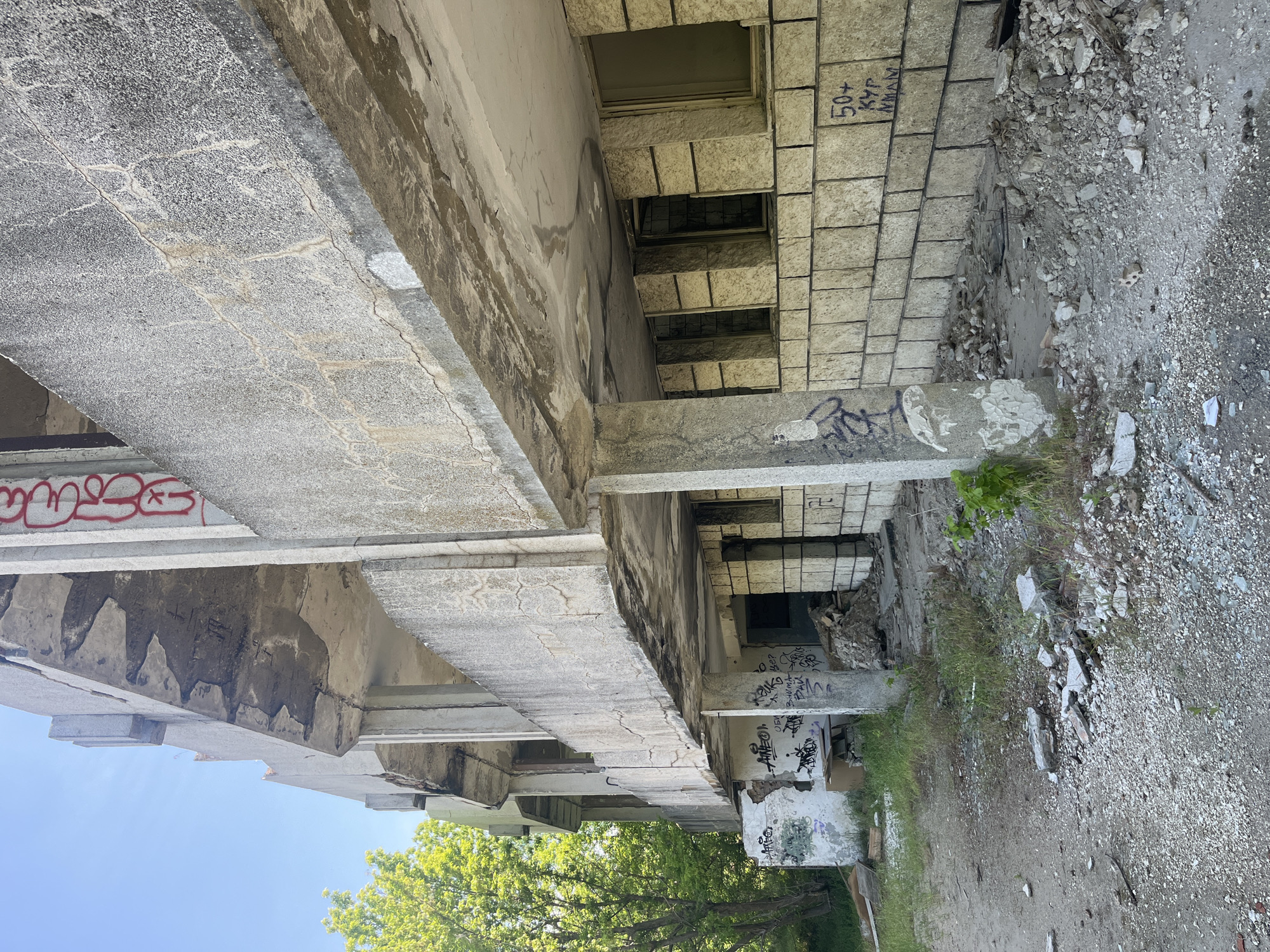 The image shows an exterior view of a derelict concrete building, possibly abandoned or under construction, in Varna, Bulgaria. The architecture features exposed concrete beams, columns, and walls. Graffiti is visible on the structures. Windows are boarded up, and there is evidence of decay and debris scattered around. The environment includes vegetation growing near the building's base, gravel, and soil. The sky is visible in the background. There are no people evident in the image.