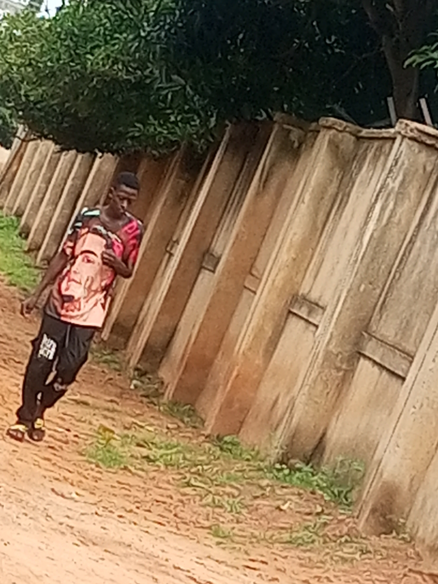 A young Black male walks along a dirt path next to a tall, weathered concrete wall. The man is wearing a pink t-shirt with a large portrait of a face printed on it, and black pants with a white logo. He is looking down at his shirt, and has his hand to his mouth. The wall is made of large, vertical concrete slabs, and there is some green foliage growing along the base of the wall. The ground is a mixture of dirt and sparse grass. The scene is outdoors, and the lighting suggests it is daytime.