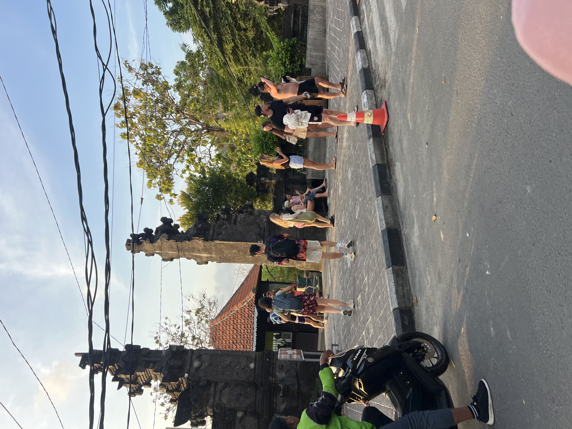 This image captures a scene in front of a traditional Balinese stone gate in Kuta Selatan, Indonesia. Several people are present. On the left, a person wearing a neon green jacket and dark pants is seated on a black scooter. To the right of the gate, a group of tourists are gathered. Some are standing, while others are sitting on a low stone wall. They appear to be diverse in age and gender, dressed casually in shorts, t-shirts, and sundresses. One person in the group is wearing a backpack. In the foreground, a red traffic cone is visible on the paved area. Beyond the gate, a building with a terracotta tile roof can be seen, and lush green trees form the backdrop. Overhead, power lines crisscross the sky, which is a pale blue with some wispy clouds. The sun appears to be setting, casting long shadows and creating a warm, golden light.