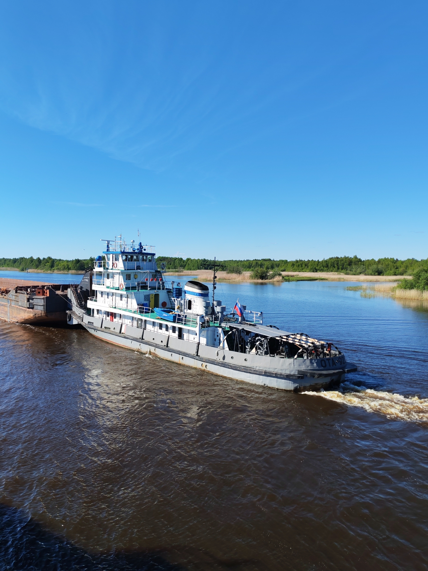 A large grey and white tugboat is actively pushing a dark-colored cargo barge along a wide, brown-hued river under a bright, clear blue sky. The tugboat is positioned at the stern of the barge, generating a significant foamy wake as it propels the vessels forward. Lush green trees and reeds densely line the distant river banks on both sides, with the vegetation extending to the water's edge.
The sky is largely cloudless, featuring only a few faint, wispy cirrus clouds. The bright illumination suggests it is daytime, likely morning or afternoon, with excellent visibility. A small Russian flag (white, blue, red horizontal stripes) is visible flying from the tugboat's superstructure.
Visible text on the tugboat includes "ОТА-92" prominently displayed on its stern. Further forward, on the superstructure, the text "Д 7786" is visible, along with a partially obscured word that appears to begin with "СУДОВОЙ" (meaning 'ship's' or 'vessel's' in Russian). This scene depicts commercial river transport in Kargulino, Russia.