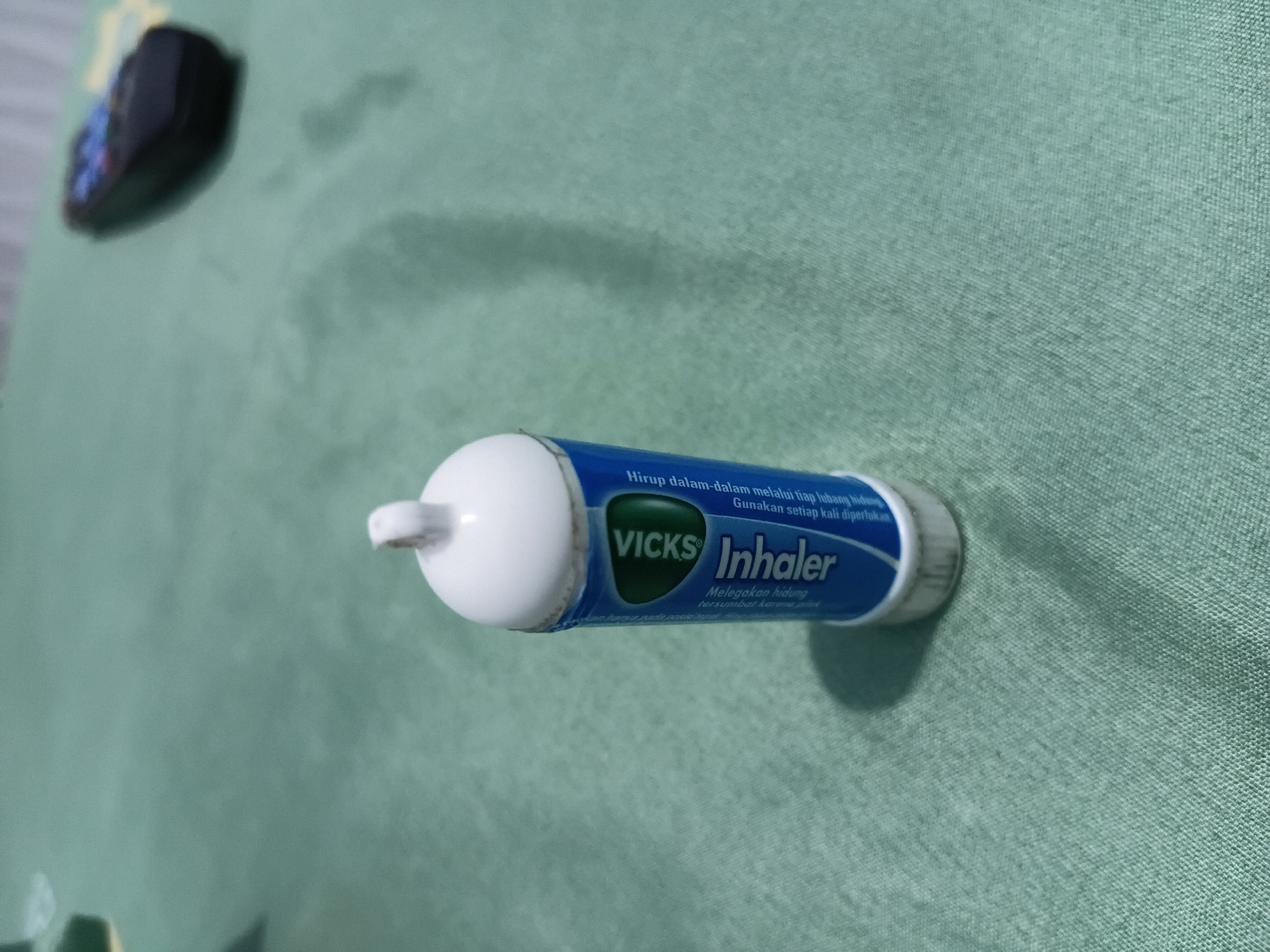 A close-up, eye-level shot shows a Vicks Inhaler resting on a green, textured surface, likely a fabric tablecloth, in what appears to be a domestic setting. The inhaler, a white cap atop a blue cylinder with white lettering, is positioned slightly off-center to the right. The text on the cylinder includes the brand name "Vicks" in a green oval and "Inhaler" in white capital letters. Some smaller text, possibly instructions or product information, is also visible in white and black. The background is a soft-focus, muted green. To the upper right of the inhaler, a blurry, dark object with colorful lights is visible, resembling a television remote control. The lighting is moderate, casting subtle shadows that suggest an indoor environment. The image does not provide any specific geographical indicators that would place it in Tanggul Wetan, Indonesia.