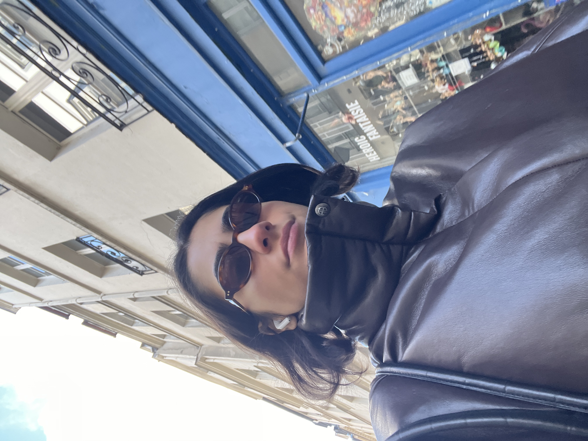 A person wearing sunglasses and a dark-colored leather jacket is taking a selfie while standing on a street in Paris, France. Buildings with traditional Parisian architecture are visible in the background. The sign "HEROIC FANTAISIE" is visible in reverse in a shop window.