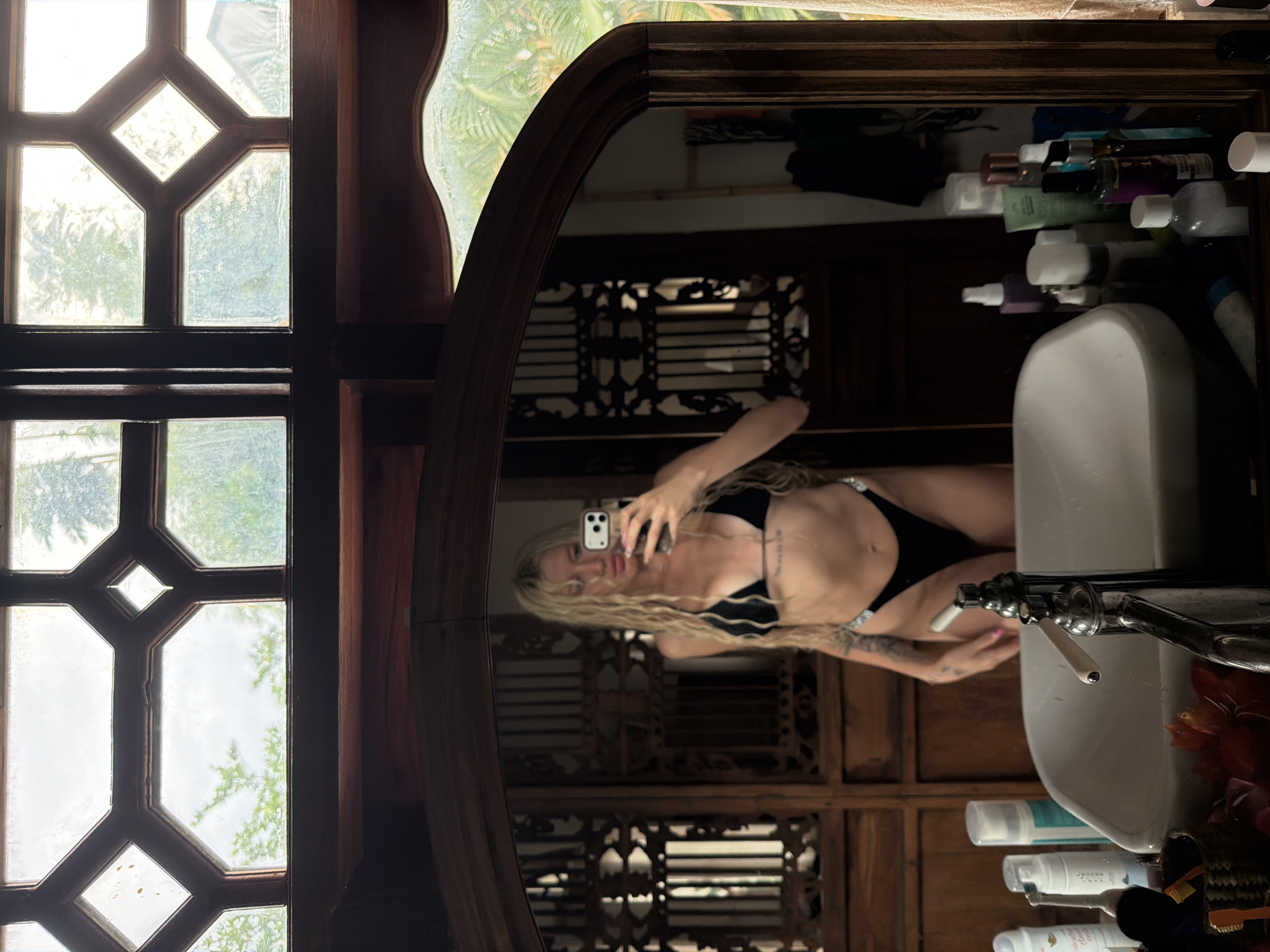 In North Kuta, Indonesia, a woman with long, blonde, wavy hair is taking a selfie in a bathroom mirror. She is wearing a black bikini with rhinestone detailing on the sides of the bottoms. The bathroom features ornate wooden paneling and a large, antique-style mirror. Through a window with a geometric wooden frame, lush green foliage and an overcast sky are visible. On the bathroom counter, a white sink is visible, along with various toiletries and a faucet. The overall ambiance suggests a tropical or exotic location.