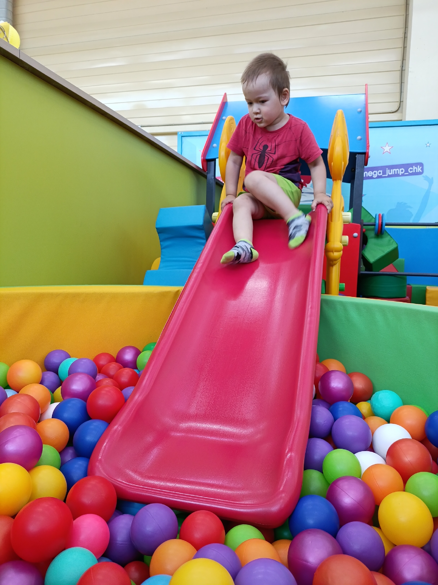 A toddler is at an indoor playground in Chaikovsky, Russia. The boy is sitting at the top of a red slide. He is wearing a red t-shirt with a spider emblem and green shorts. The slide empties into a pit filled with colorful plastic balls. The playground equipment is set against a backdrop of green and blue padded walls. "nega_jump_chk" is written on the blue wall behind the slide.