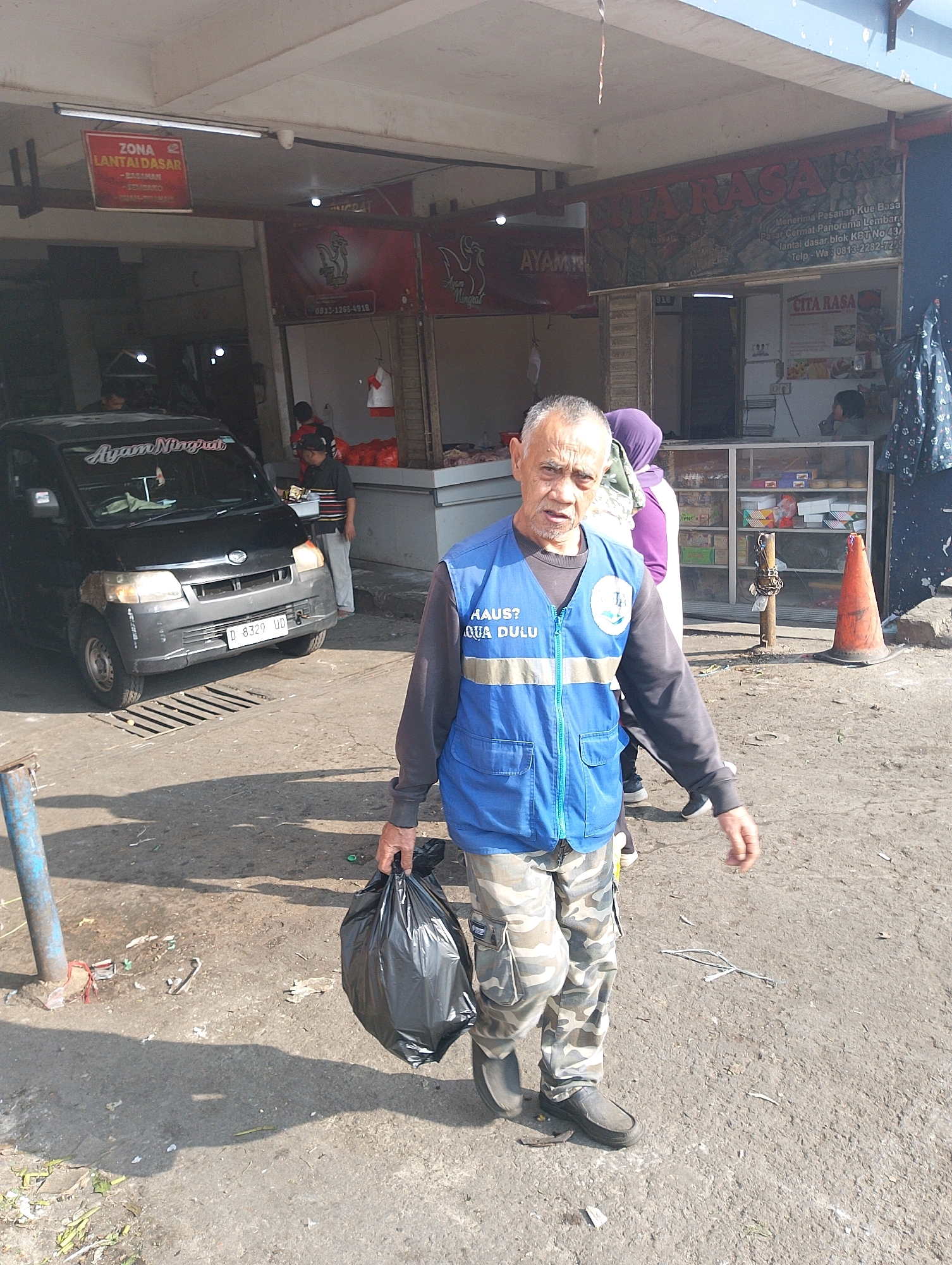 In Lembang, Indonesia, a man wearing a blue vest with reflective stripes over a gray long-sleeved shirt and camouflage pants walks towards the viewer, carrying a black garbage bag in his right hand. The man has graying hair and a weathered complexion. Behind him, a black Daihatsu van with an Indonesian license plate is parked, and to the right, a storefront displays various products. The ground is a mixture of dirt and pavement with some scattered debris. The scene appears to be an outdoor market or commercial area.