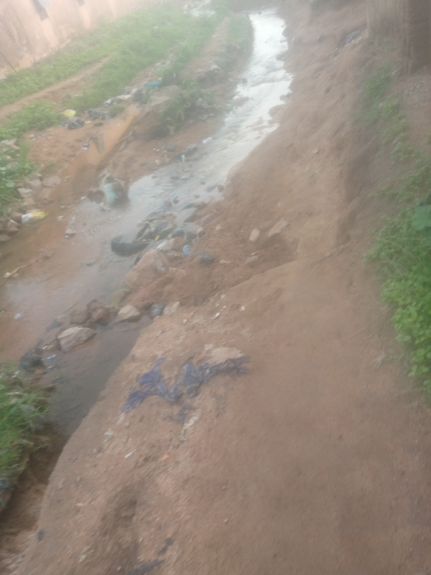 The image depicts a dirty drainage channel flowing through a predominantly unpaved area in Jalingo, Nigeria. The channel is filled with murky water and debris, including plastic waste and what appears to be discarded tires. The banks of the channel are made of earth and rocks, with some sparse vegetation growing on them. A building, possibly a wall, is visible in the background, out of focus. The overall impression is one of neglect and environmental degradation.
