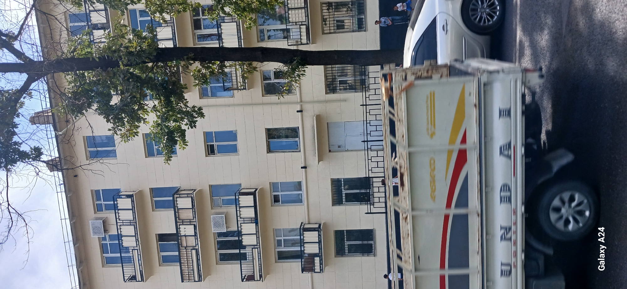 In Bishkek City, Kyrgyzstan, a light-colored, multi-story building with multiple balconies and windows is visible. The building has a cream-colored facade. Some windows are open, while others are closed. Several air conditioning units are mounted on the exterior walls. A large tree with green leaves partially obstructs the view of the building. In the foreground, the side of a white truck with colorful stripes and the word "HYUNDAI" is prominently displayed, indicating it is a Hyundai Porter model. A silver car is partially visible to the right of the truck. A black metal fence with a decorative pattern runs along the ground floor of the building. Three people are visible standing near the fence: a man in a gray shirt and blue jeans, a child in a blue shirt and shorts, and another child in a white shirt. The scene appears to be a street view on a sunny day.