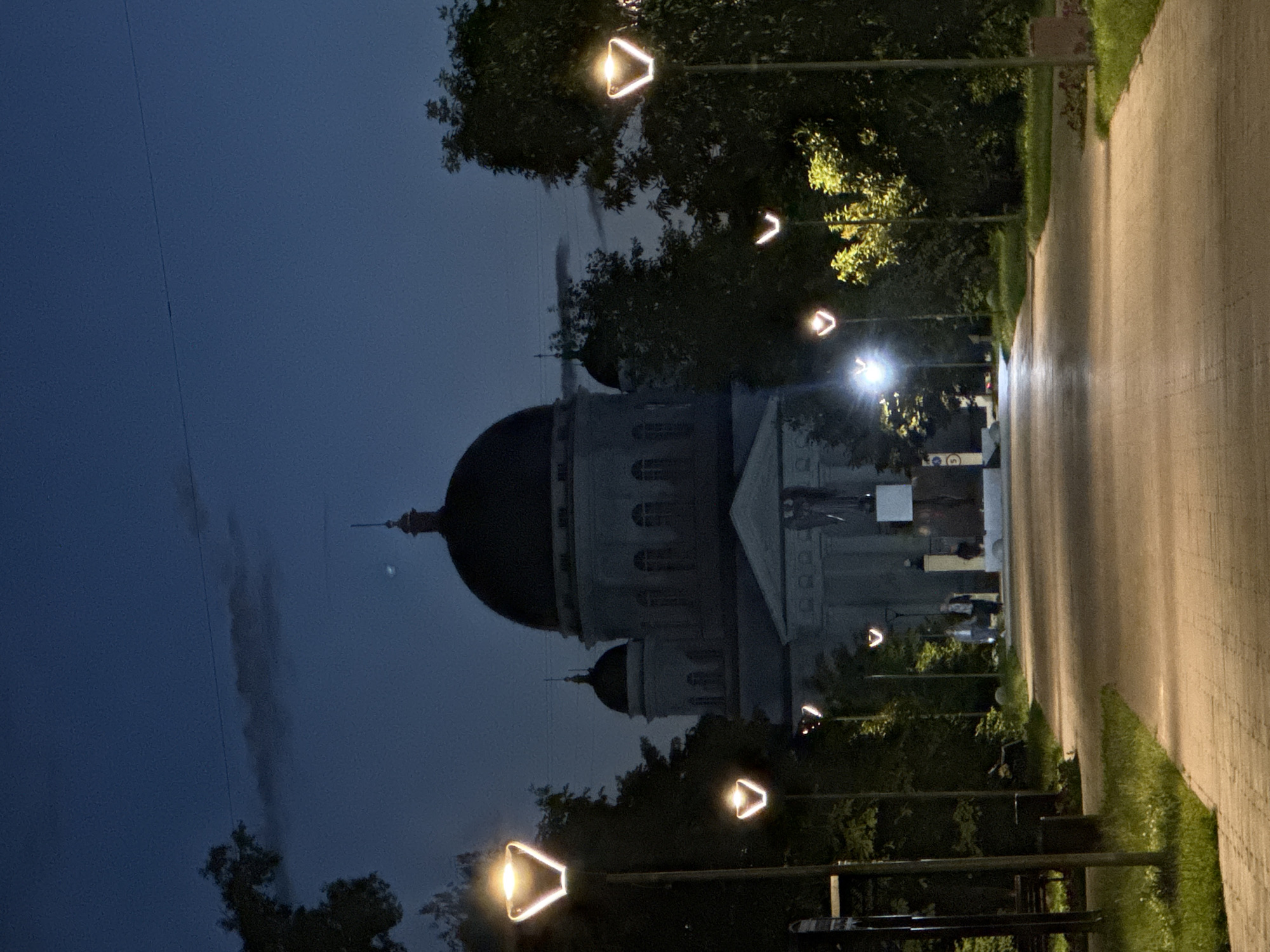 The image shows a paved walkway leading towards a large domed building at dusk or night. The walkway is lined with modern streetlights with distinctive triangular light fixtures. On the right side of the walkway, there are trees and bushes. On the left side, there are also trees, grass, a bench, and a trash can. In the background, the domed building, possibly a church or government building, is dimly lit, with smaller domed structures attached. There are a few people walking on the walkway in the mid-ground. The sky is dark blue with some wispy clouds and a faint light source visible in the upper right portion of the sky. The overall scene suggests a public park or square area in a city. This location is identified as the Cathedral of Alexander Nevsky in Nizhny Novgorod, Russia.