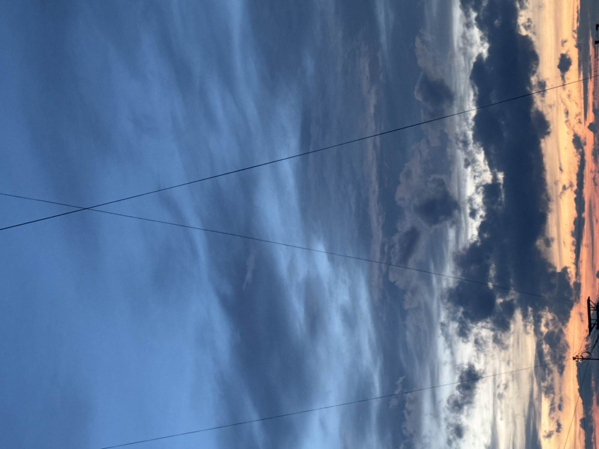 This media file is a wide-angle photograph of a sky scene, likely captured at sunset or sunrise, with the provided context placing it in Chaikovsky, Russia. The sky exhibits a striking gradient of colors and cloud types, divided roughly into three visual sections.
On the left, the sky is a lighter, clear blue with soft, wispy clouds. Towards the center, the blue deepens, and more defined, streaky white clouds are visible, stretching across the frame. The right side of the image presents a dramatic contrast, featuring large, dark, dense clouds silhouetted against a vibrant horizon. This horizon glows with warm hues of orange, pink, and yellow, strongly indicative of either sunrise or sunset.
Several thin, dark lines, resembling utility or power lines, traverse the sky. Some run horizontally across the entire width of the image at varying heights, while others intersect diagonally in the upper-left portion. In the far right, faint silhouettes of distant structures, possibly industrial elements or a crane, can be discerned against the colorful backdrop.
There are no people or visible text in the image. The weather appears to be partly cloudy with significant cloud formations, especially on the right. The scene captures the natural beauty and expansive quality of the sky at dawn or dusk.