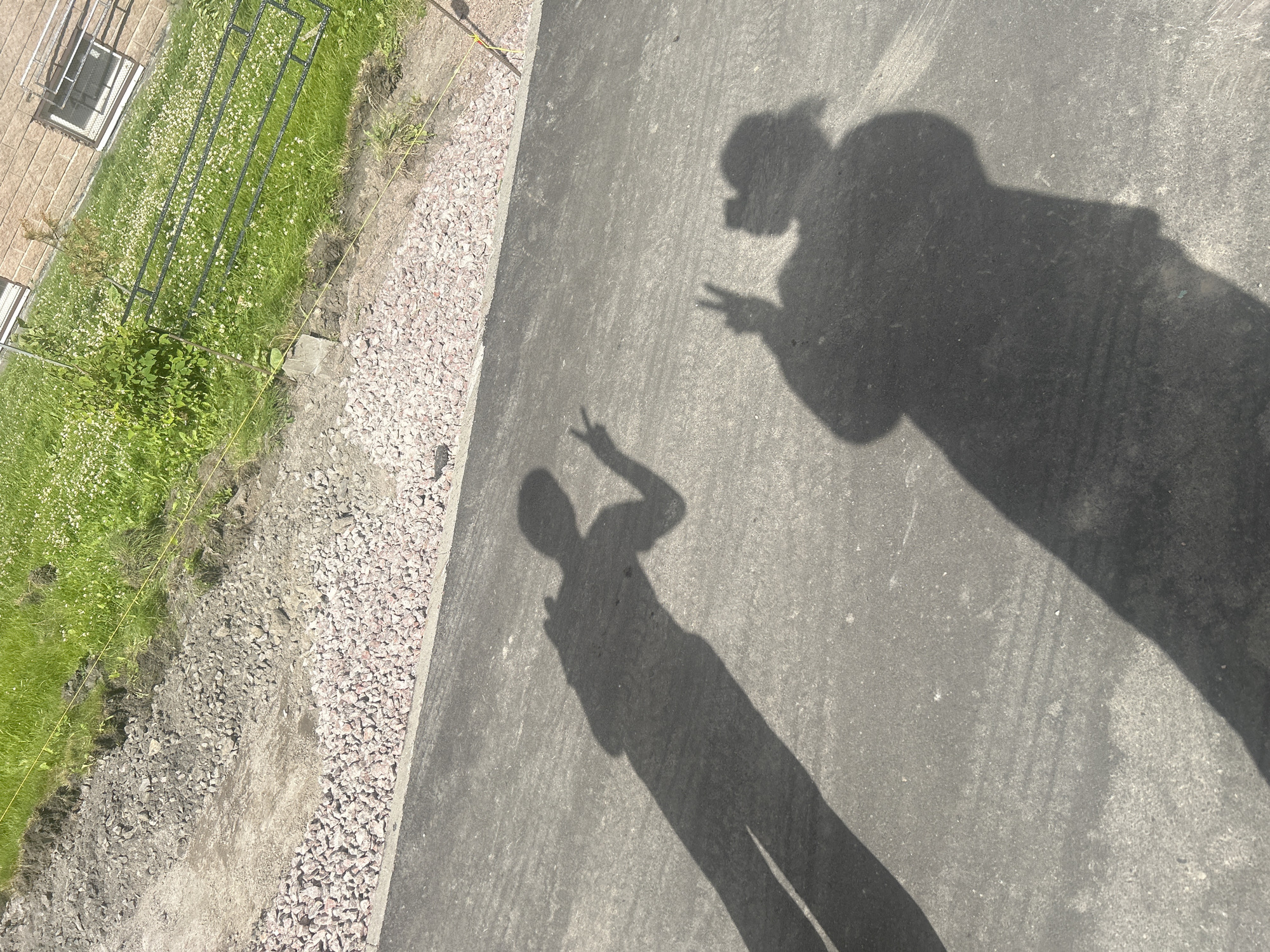 The image displays the shadows of two individuals on an asphalt surface. The individual on the left casts a shadow of a person standing with their arms extended, one hand forming a peace sign. The individual on the right casts a shadow of a person holding their hand up with a peace sign. In the background, there is a strip of pink gravel, followed by dirt and then a grassy area with white flowers. A building with brick siding and windows is visible in the upper right corner. A black metal fence runs through the grassy area, and a yellow string or wire is stretched horizontally across the scene. The lighting suggests it is daytime, likely a sunny day given the distinct shadows. This scene appears to be a construction or landscaping area, possibly near a residential building, in an outdoor urban environment. The specific location in Saint Petersburg, Russia, cannot be determined from the visual information provided.