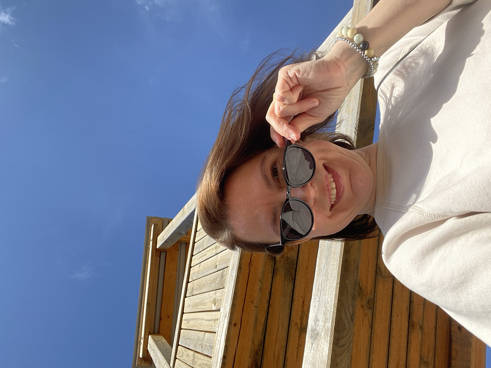 A woman with shoulder-length brown hair is shown from a low-angle perspective, smiling and adjusting her sunglasses. She is wearing a light-colored sweatshirt and two beaded bracelets on her left wrist. Behind her, a wooden structure with slats and beams is visible, and beyond that, a clear blue sky with wisps of clouds. The photo appears to be taken outdoors. No information about the location in the city of Chaikovsky, Russia can be discerned from the image.