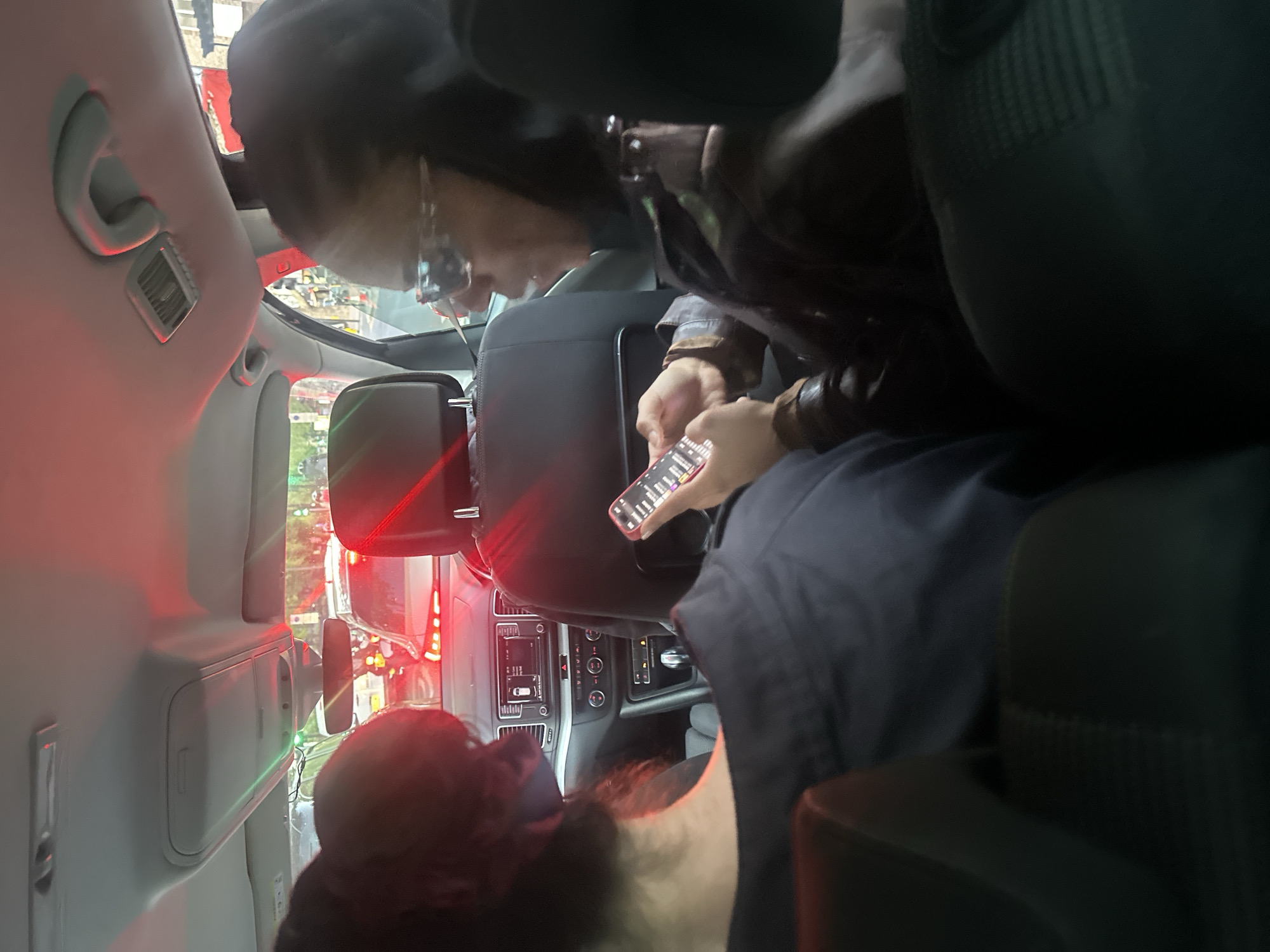 The image depicts the interior of a passenger vehicle, likely an SUV or van, at night or during dusk, while stationary in urban traffic. The view is from a rear seat, looking towards the front passenger side and dashboard.
In the mid-ground, a person is seated, appearing to be a passenger in the rear seat on the right side of the vehicle (when viewed from the front). They are wearing a dark top, possibly a hoodie, and glasses. Their head is bowed, focusing intently on a red smartphone held with both hands. The phone screen is brightly illuminated, displaying an interface with several indistinct icons and text elements that are not clearly legible.
The vehicle's interior features a light-colored headliner with grey grab handles and air vents. The dashboard is visible towards the front, housing a central console with various controls including a radio/infotainment screen, climate controls, and buttons such as the hazard lights. A rear-view mirror reflects some external lights and a partial figure, possibly the driver.
Through the front and side windows, the exterior scene reveals heavy urban traffic. Dominant red lights from brake lights or taillights of vehicles ahead create a strong red glow across the scene, indicating congestion. Green and white lights from streetlights or other vehicles are also visible in the distance. The setting suggests a busy street in London, United Kingdom. No specific text is clearly decipherable on the phone screen or car dashboard controls.