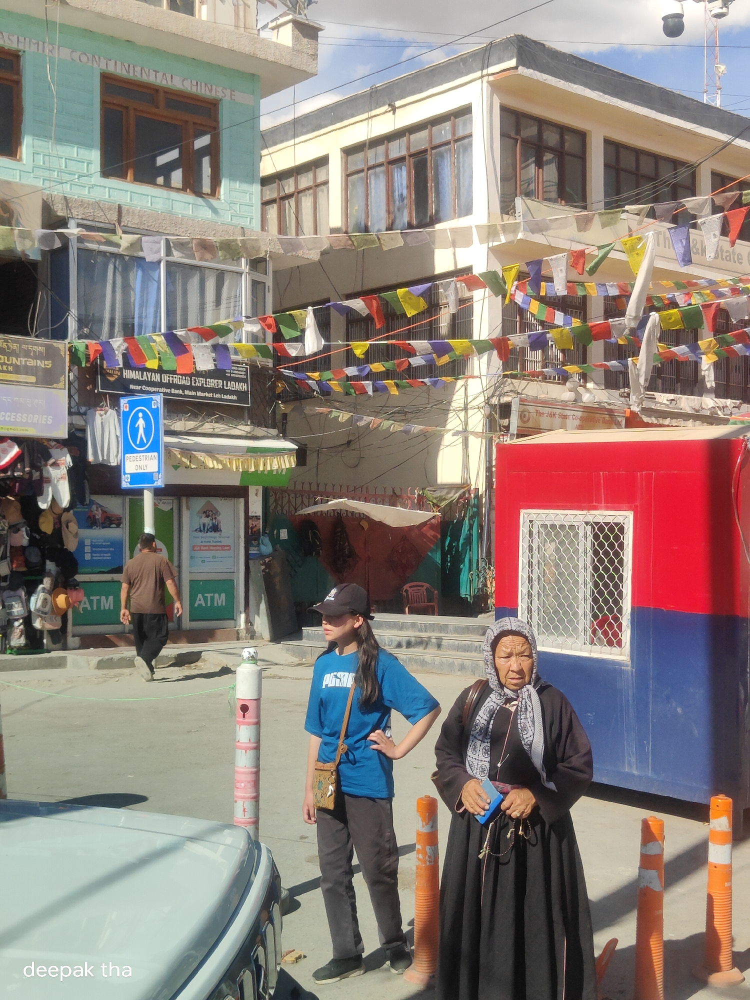 The image depicts a street scene in Leh, India, under a bright, partly cloudy sky. In the foreground, a white vehicle is partially visible on the left. To the right of the vehicle, a young person wearing a blue t-shirt and dark pants stands with one hand on their hip. Next to them, an elderly woman, dressed in a black long-sleeved garment and a patterned headscarf, stands facing the camera, holding prayer beads and a small blue object.
Behind the people, the street is lined with multi-story buildings adorned with strings of colorful prayer flags. The building on the far left has a light blue facade with "KASHMIR CONTINENTAL CHINESE" written in white lettering. A pedestrian crossing sign with a blue background and a white silhouette of a person is prominently displayed on a pole. Several ATM machines are visible through glass storefronts.
In the middle ground, a man in a brown t-shirt and dark pants walks away from the camera. A bright red and blue structure, possibly a kiosk or small building, is situated on the right side of the frame. Various shops and market stalls with merchandise are visible on the left. The overall environment suggests a busy commercial street in a mountainous region.