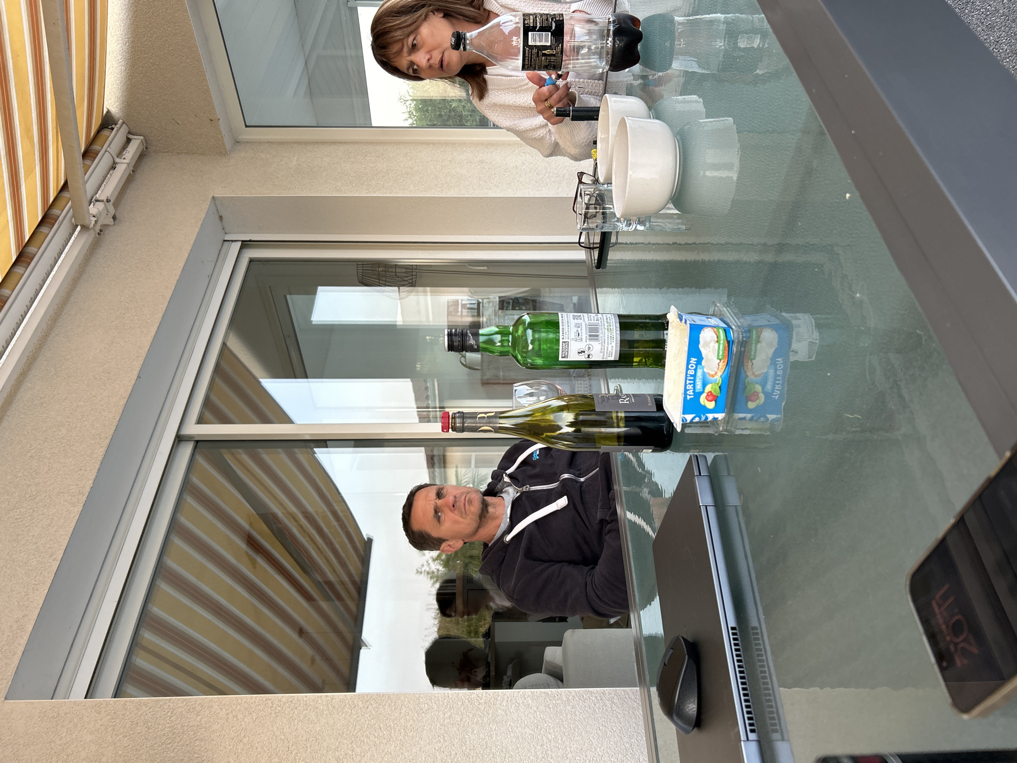 Here is a detailed description of the image:
Two adults are seated at a glass-topped outdoor table. One adult has brown hair and is wearing a white sweater. They are holding a water bottle. The second adult is a man with dark hair wearing a black jacket and a gray t-shirt.
On the table are a wine bottle, a green bottle, a package of cheese, two white bowls, a water bottle, glasses, a laptop, a mouse, and a smartphone.
The scene appears to be on a patio. There is a window and a striped awning in the background.