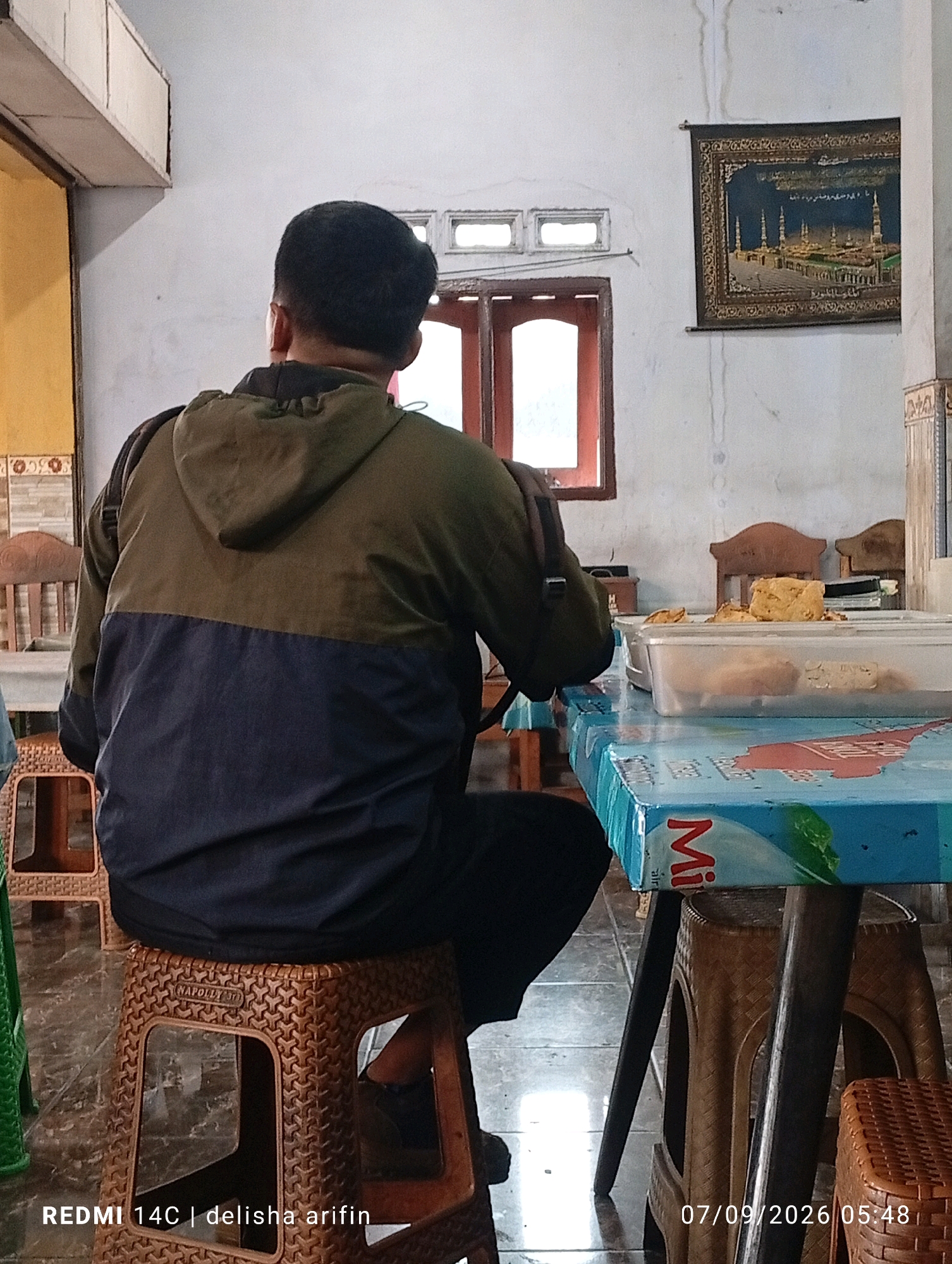 A rear view of a man in a two-toned green and navy hooded jacket is captured, seated on a brown woven plastic stool. He appears to be looking towards a table covered with a colorful, patterned tablecloth. On the table, several containers of food are visible, including a clear plastic tub filled with what looks like fried snacks. The scene is set indoors, with a white wall and a framed tapestry depicting a mosque in the background. Wooden chairs and windows with red frames are also present in the environment. The timestamp "07/09/2026 05:48" and the text "REDMI 14C | delisha arifin" are overlaid on the bottom right and left corners of the image, respectively. The overall impression is of a casual meal being served or consumed in what appears to be a modest dining area.