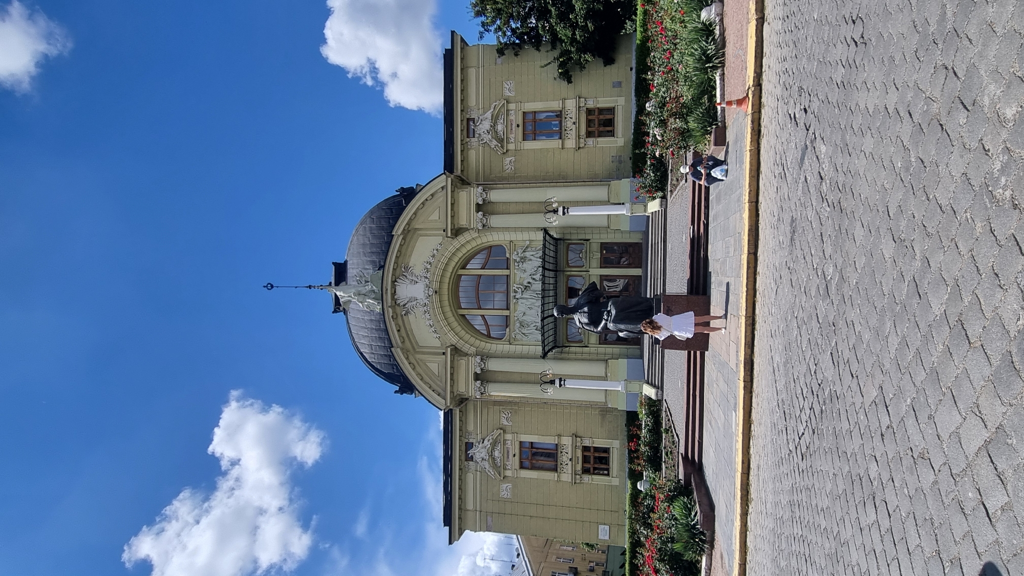The image shows the Chernivtsi Musical Drama Theatre, also known as the Olha Kobylianska Chernivtsi Regional Ukrainian Music and Drama Theatre, in Chernivtsi, Ukraine. The theater is a prominent architectural landmark with a distinctive dome. In front of the theater, there is a bronze statue of a seated figure. A young woman in a white dress stands facing the statue, while a person wearing a baseball cap and dark clothing is seated on the ground to the right, with a plastic bag and an orange traffic cone nearby. The area is paved with cobblestones, and there are flower beds with red roses in front of the building. The sky is blue with scattered white clouds.
