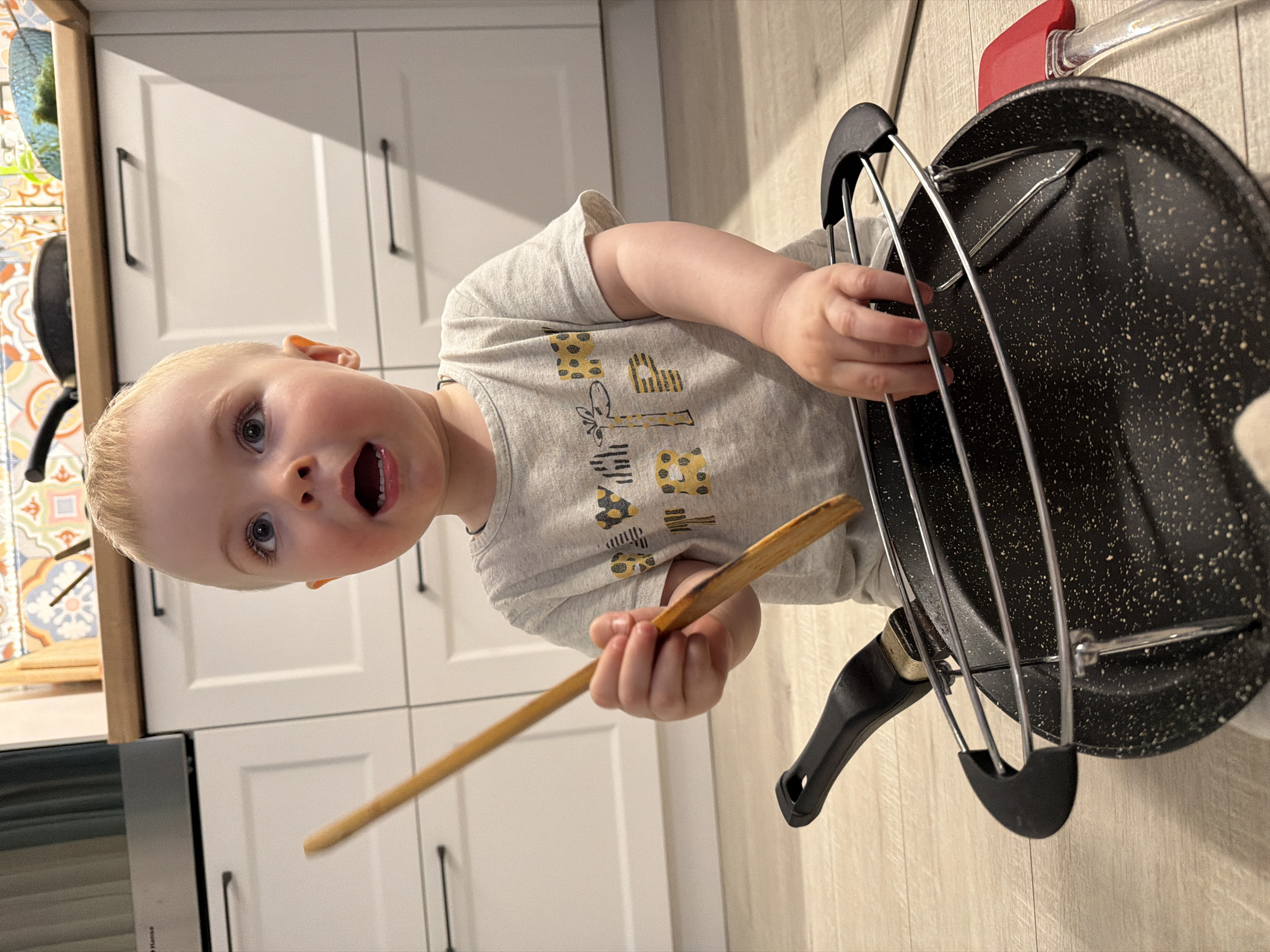 A blonde toddler stands in a kitchen, holding a wooden spoon in one hand and touching a pan rack with the other. The kitchen features white cabinets with black handles, a tiled backsplash, and a stainless steel oven. The location is not specified, and there is no indication of it being in Votkinsk, Russia.