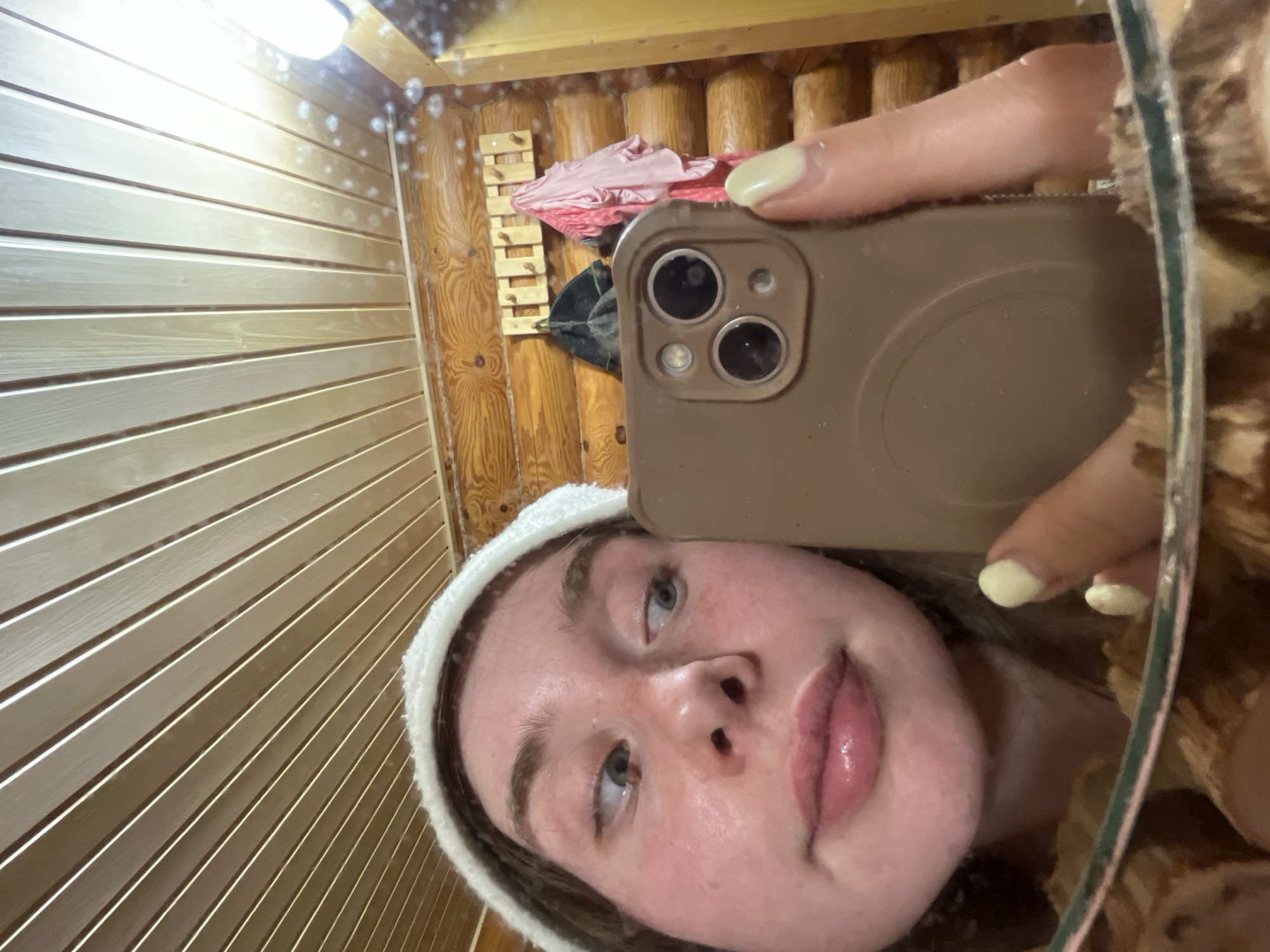 A selfie of a woman with fair skin and blonde hair, wearing a white headband, is reflected in a mirror. She is holding a brown smartphone with multiple camera lenses, taking the photo. The mirror is surrounded by a wooden frame, reflecting a room with wooden walls and a clothing rack holding pink and black items. The left side of the image shows a wall made of light-colored horizontal planks. The location is likely indoors and based on the request, is inferred to be in Chaikovsky, Russia.