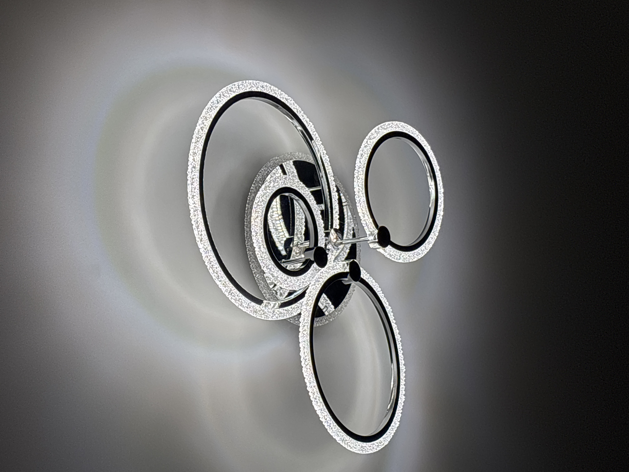 A modern, multi-ringed ceiling light fixture is depicted against a light gray ceiling, transitioning to a darker gray towards the bottom of the frame. The fixture consists of several interconnected, elliptical, and circular rings, varying in size and orientation, each outlined with numerous small, glittering elements that emit a bright, diffuse light. The inner surfaces of the rings appear dark, contrasting with the illuminated outer edges. The central mounting point of the fixture is visible, featuring a reflective, metallic surface and several attachment points. The light from the fixture casts subtle, circular light patterns and soft shadows on the surrounding ceiling. A faint, arc-shaped spectrum of colors, indicative of light diffraction, is noticeable on the ceiling above and to the right of the fixture. The image is captured from a low-angle perspective, looking directly upwards at the ceiling fixture. The location is an indoor setting in Unknown, Unknown.