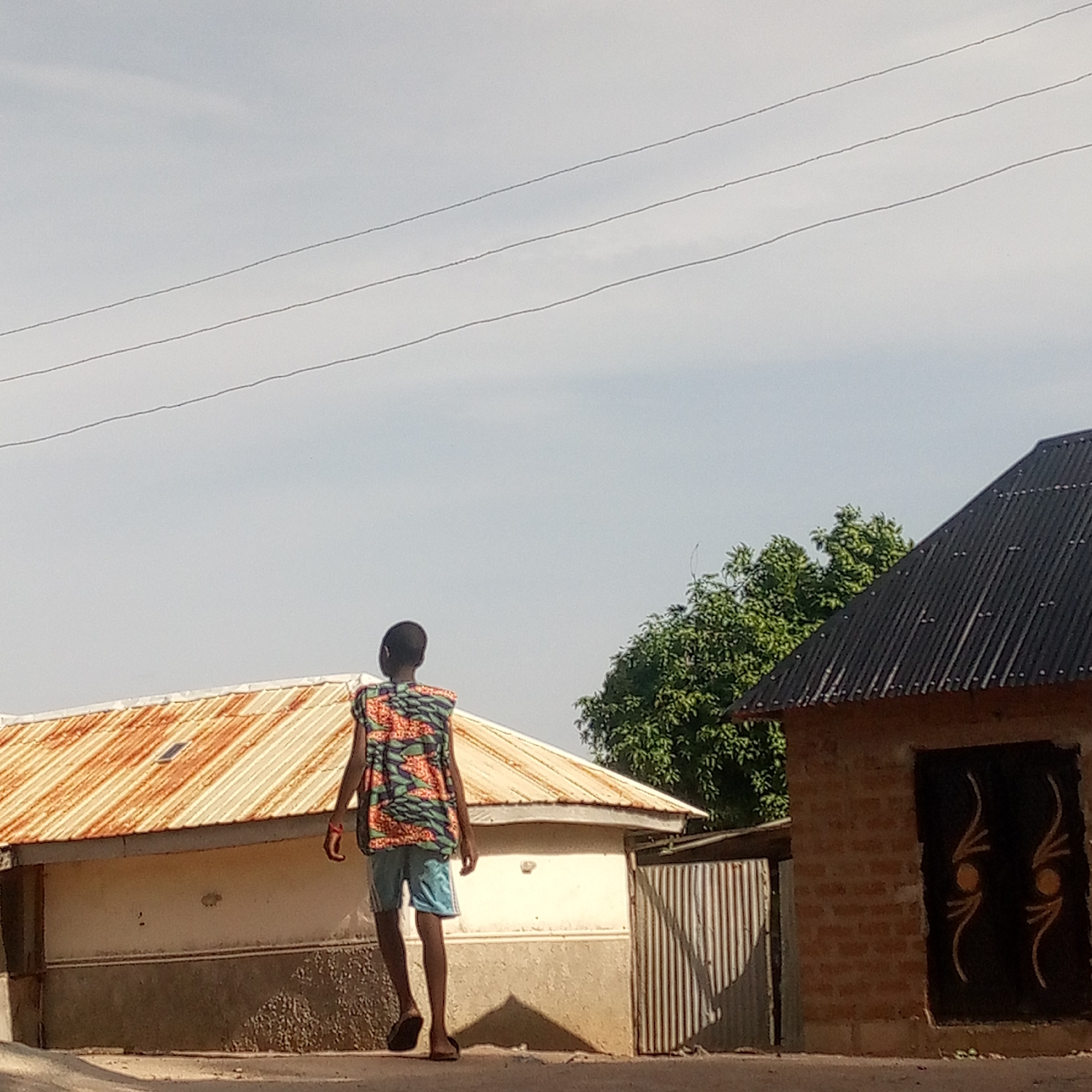 A young individual walks away from the camera in a street in Mararaba, Nigeria. The person is wearing a sleeveless, patterned top and blue shorts with flip-flops. They are walking on a paved area adjacent to two buildings. The building on the left has a rusty metal roof and a white wall with a dark lower section. The building on the right has a dark, corrugated metal roof and a brick wall with a dark door decorated with light-colored designs. There are overhead electrical wires above. A tree with green foliage is visible behind the right building. The sky is partly cloudy.