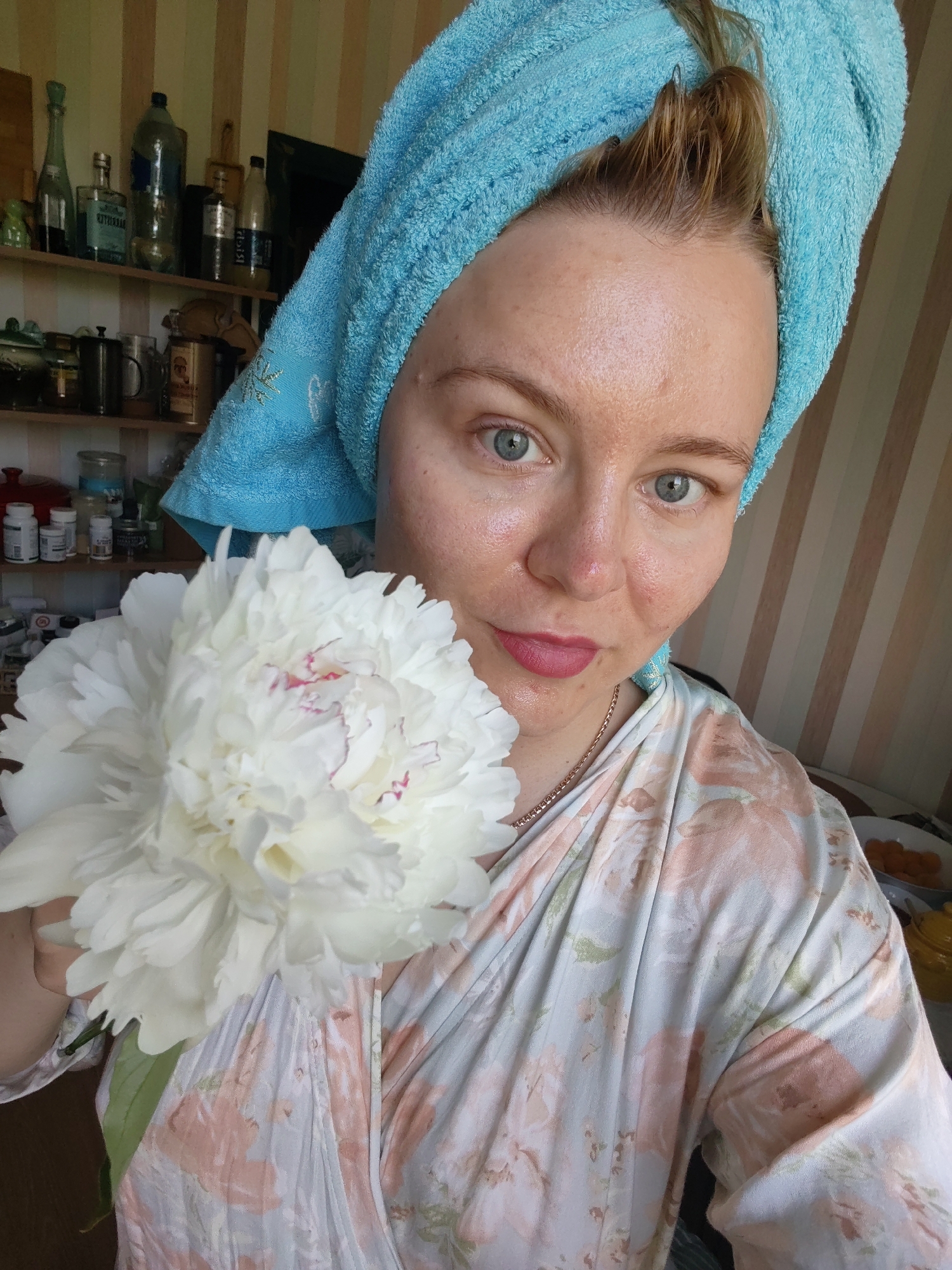 A woman with blue eyes, a blue towel wrapped around her head, and a floral robe is holding a white peony flower. She is looking at the camera, and her face is partially illuminated by sunlight. Behind her, shelves are visible with various items, including bottles and jars. The room appears to be a domestic setting, possibly a kitchen or bathroom, with striped wallpaper in the background. No specific location in Izhevsk, Russia, can be determined from the image.