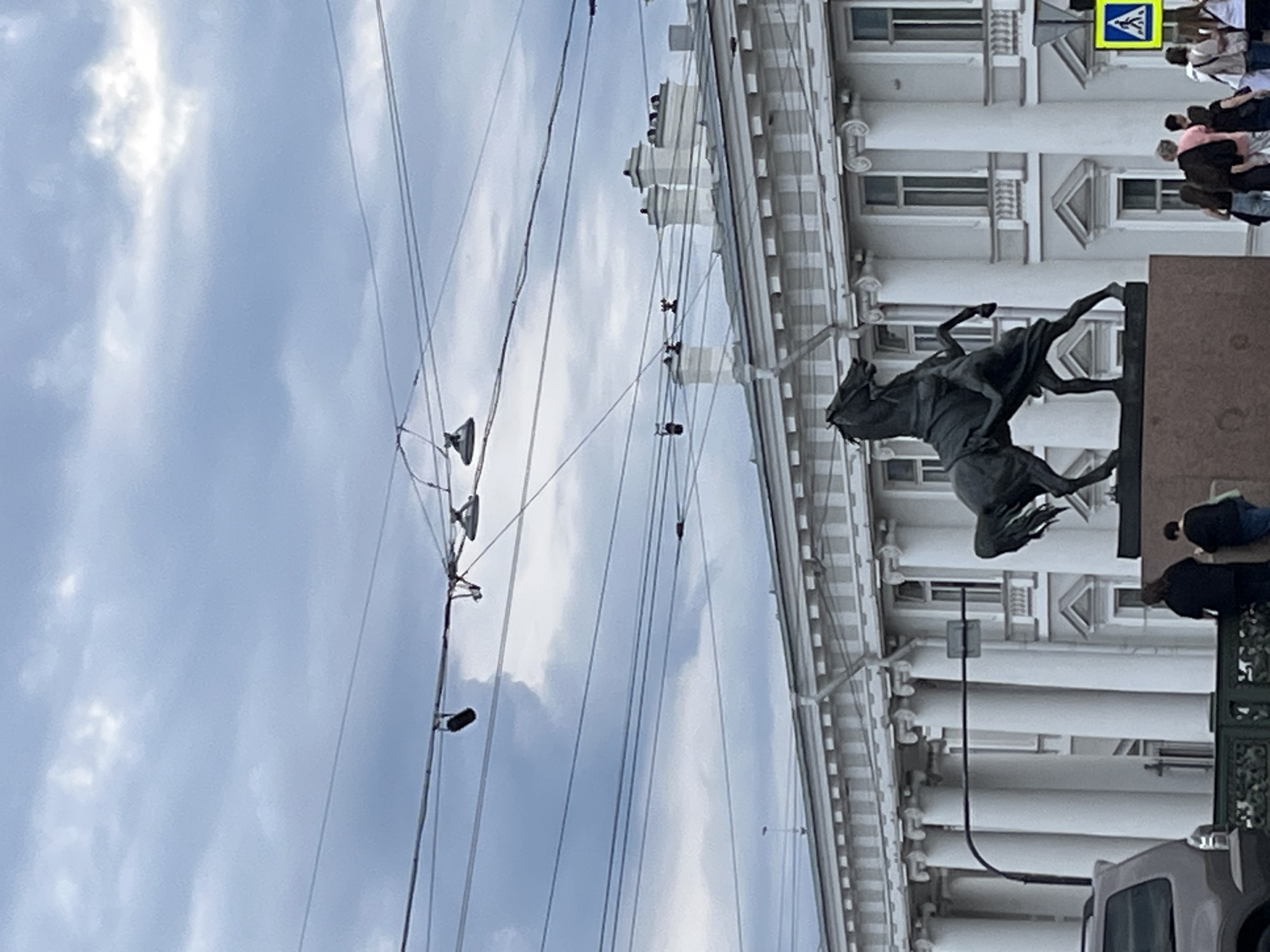 This image captures a street scene in Saint Petersburg, Russia, specifically on or near the Anichkov Bridge. In the lower-middle of the frame, a dark bronze equestrian statue, one of the famous "Horse Tamers" by Peter Klodt, depicts a man wrestling with a rearing horse on a reddish-brown granite plinth.
Behind the statue, the elegant, light-colored facade of the Beloselsky-Belozersky Palace extends across the midground. This neoclassical building features a prominent colonnade of tall white columns, regularly spaced windows, and decorative architectural details along its roofline, including several white chimneys.
The sky above is overcast, filled with various shades of light grey clouds. Numerous black electrical wires, consistent with tram or trolleybus infrastructure, crisscross horizontally and diagonally across the upper portion of the image. Several streetlights are suspended from these overhead lines.
In the lower right, a group of pedestrians, casually dressed, congregates on a sidewalk, some appearing to observe the surroundings. A yellow pedestrian crossing sign with a black human figure is mounted on a pole near this group. In the lower left, the rear section of a dark-colored SUV is partially visible. A dark, ornate metal railing runs across the bottom foreground, separating the viewer's immediate space from the street or bridge area.