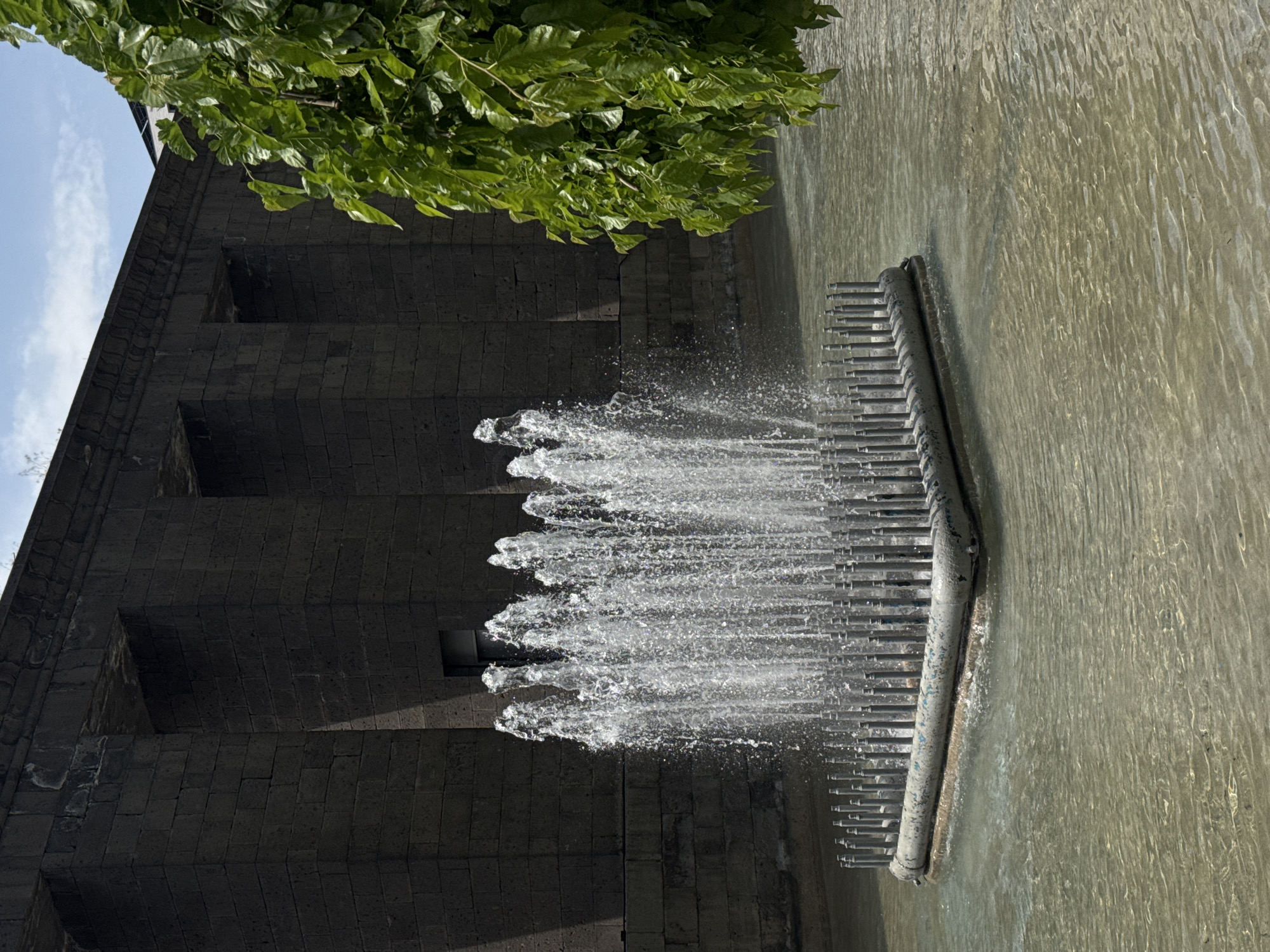 A vertically oriented medium shot shows a fountain cascading from a stone structure in Yerevan, Armenia. The fountain features horizontal metal pipes releasing water into a pool. The stone structure has evenly spaced square indentations, giving it a textured appearance. Green foliage is visible in the upper left corner. The sky is partially visible in the upper left as well.
