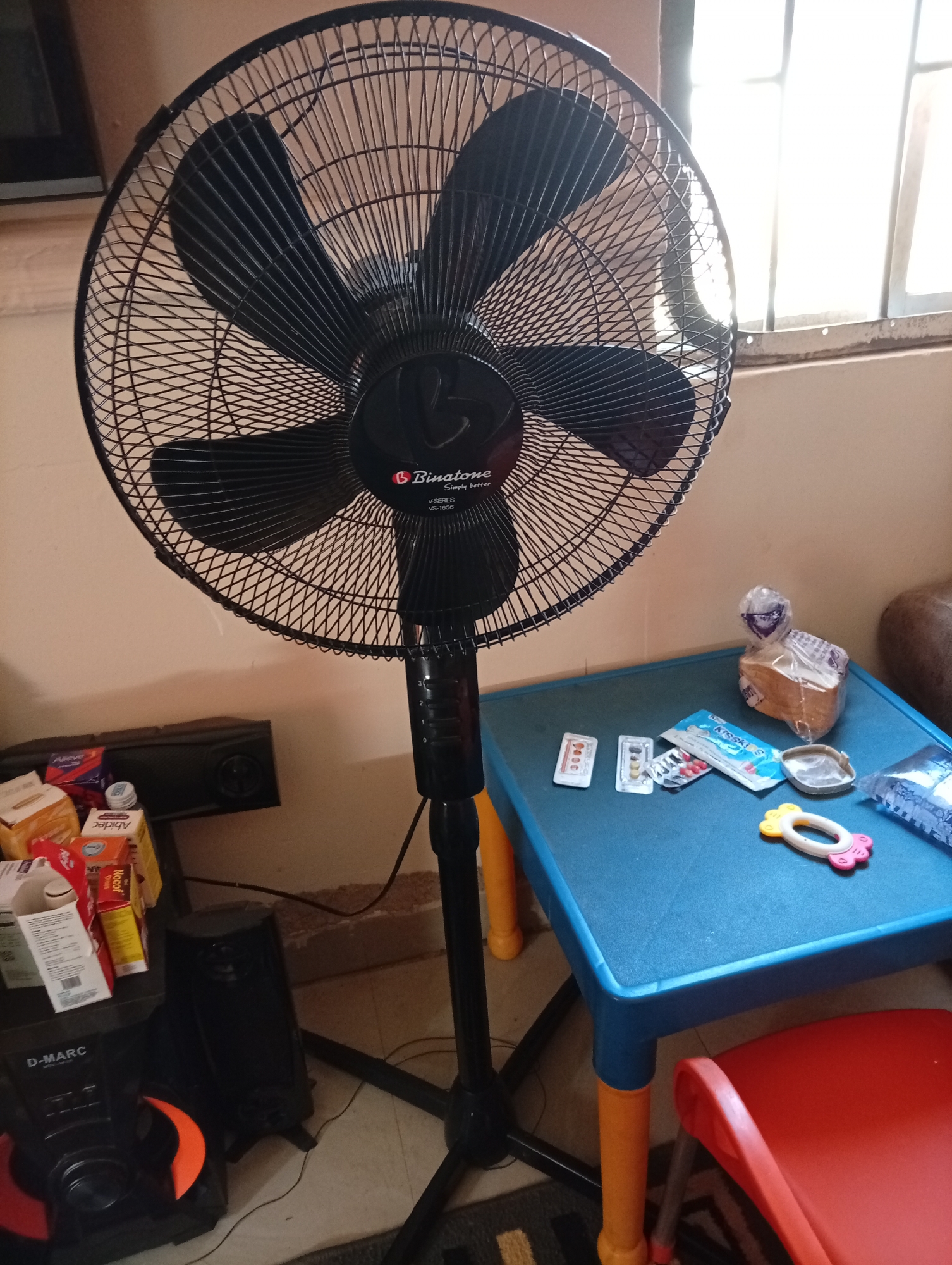 In Gura Top, Nigeria, a Binatone pedestal fan stands in a room, its blades dark and motionless. To the right of the fan, a small blue table with orange legs displays various items: a half-eaten loaf of bread wrapped in plastic, blister packs of medication, a packet of "Kisskids" candies, and a small, yellow and pink baby teether. A red plastic chair is positioned beneath the table. To the left of the fan, a black speaker system with "D-MARC" branding is visible, along with stacks of boxes and packages on a surface. The background shows a cream-colored wall and a window with a grid pattern. The lighting in the room is natural, coming from the window.