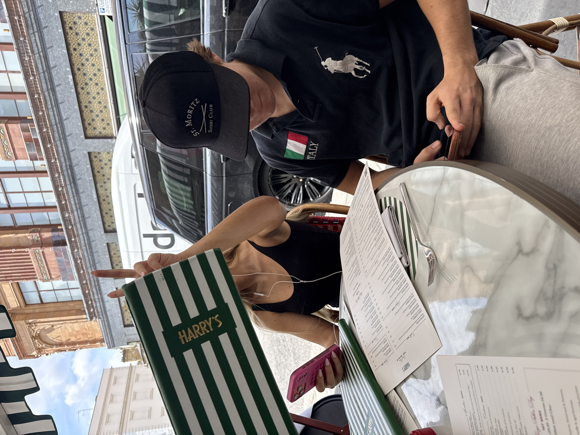 A blonde woman and a young man are seated at a marble-topped outdoor table in front of a building with ornate architecture. The woman is holding a pink smartphone and wearing a black tank top and earbuds. The man is wearing a black polo shirt with an Italian flag patch and a black baseball cap, and he is looking at his phone. A menu from "Harry's" with green and white stripes is visible on the table. The background shows a building facade, a black SUV, and a white van. The scene appears to be from a restaurant or cafe in London, United Kingdom, likely near a busy street given the presence of vehicles and the architecture.