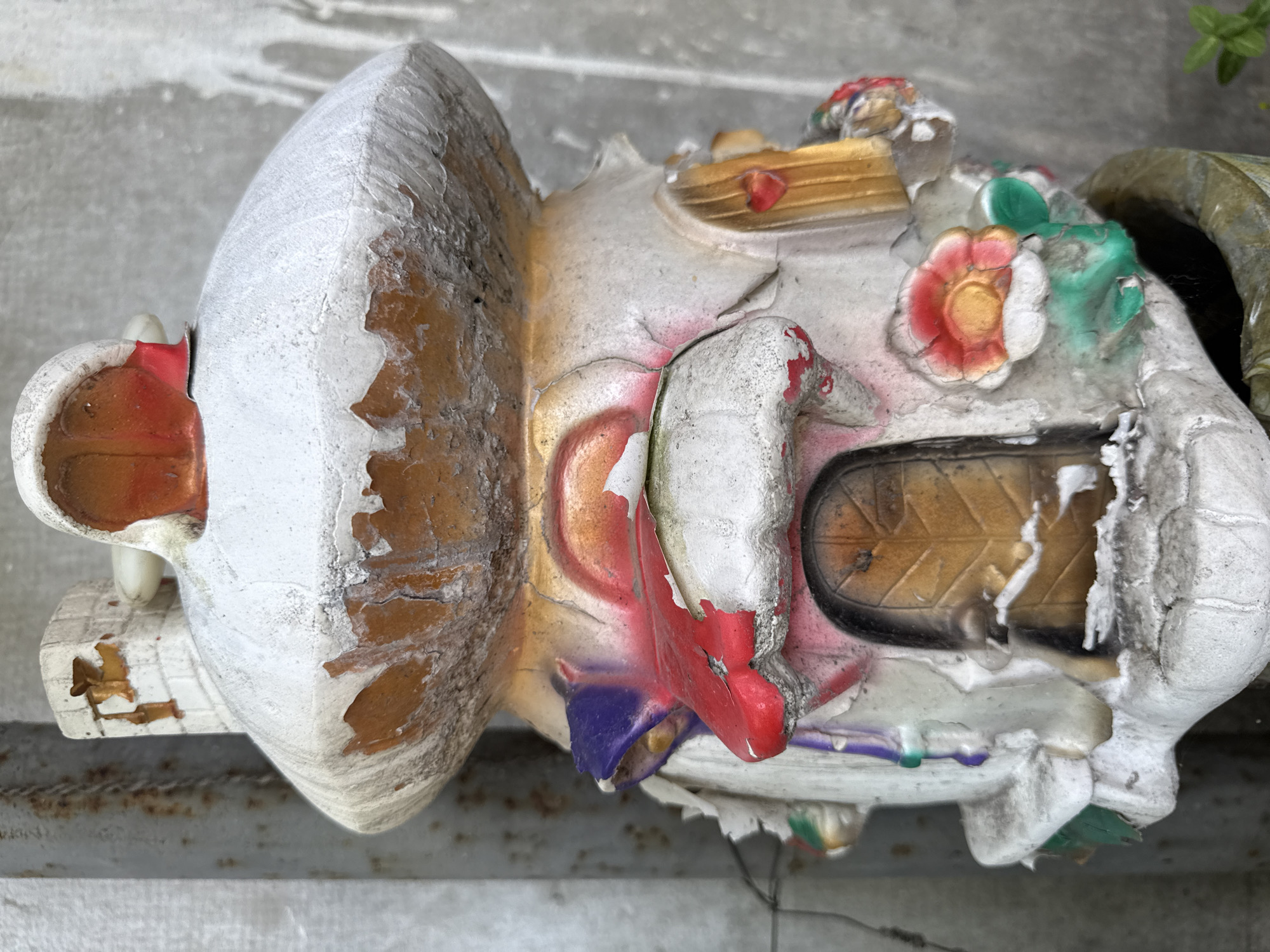 An aged, whimsical gnome or fairy house statue stands in an outdoor setting. The statue, primarily white with accents of red, orange, yellow, blue, and green, depicts a mushroom-shaped dwelling with multiple doors, windows, and floral embellishments. The paint is peeling and chipped, revealing an orange-brown material underneath, indicating significant wear and age. There is a chimney on the left side of the structure. The background consists of a textured gray concrete wall and a vertical metal pole with rust marks. The statue is placed in a pot, partially visible at the bottom of the frame.