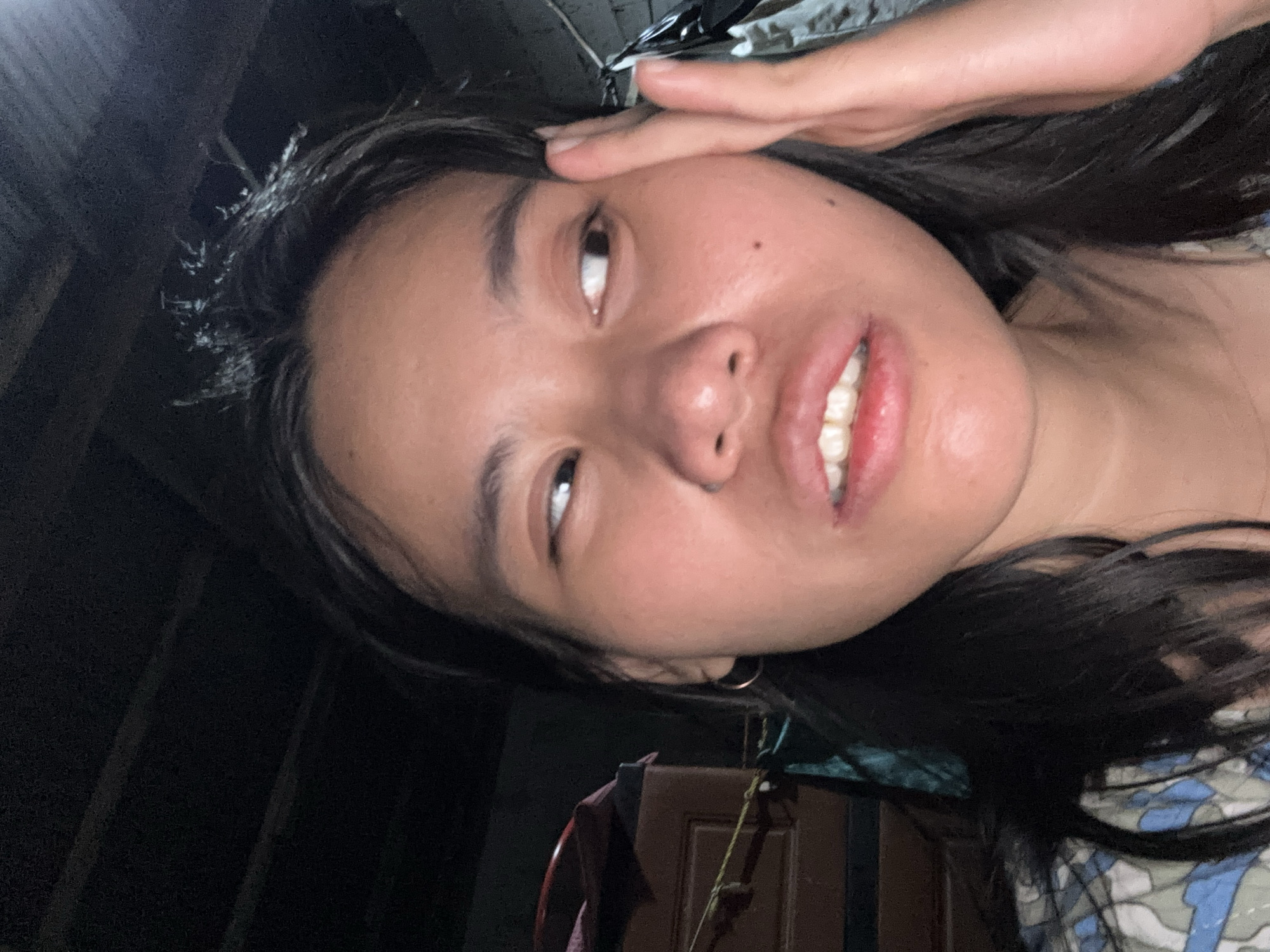 The image shows a close-up of a young East Asian woman's face, taken indoors. She has dark hair and is looking off to her right with her eyes wide. Her hand is raised to her forehead, and her mouth is slightly open, revealing her teeth. The background is dark and appears to be a room with some wooden furniture visible on the left and a wall on the right. The lighting suggests it is nighttime or a poorly lit indoor space. The specific location in East Jakarta, Indonesia, is not identifiable from the visual information provided in the image.