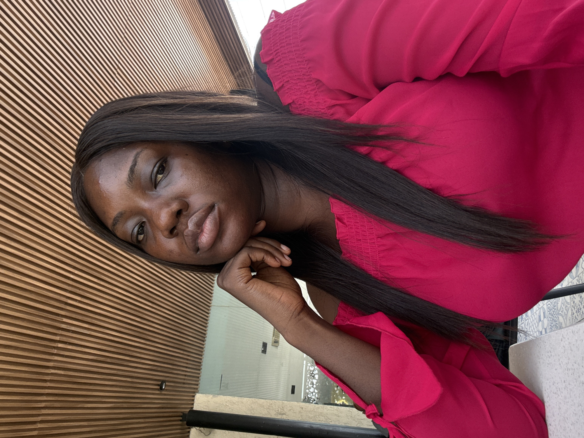A medium-angle indoor shot shows a dark-skinned woman in a red top. She leans her head on her left hand, facing the camera. Her long, dark hair is straight. The background features a wall with horizontal wooden slats and a partial view of a window. The location is likely within Abuja, Nigeria.