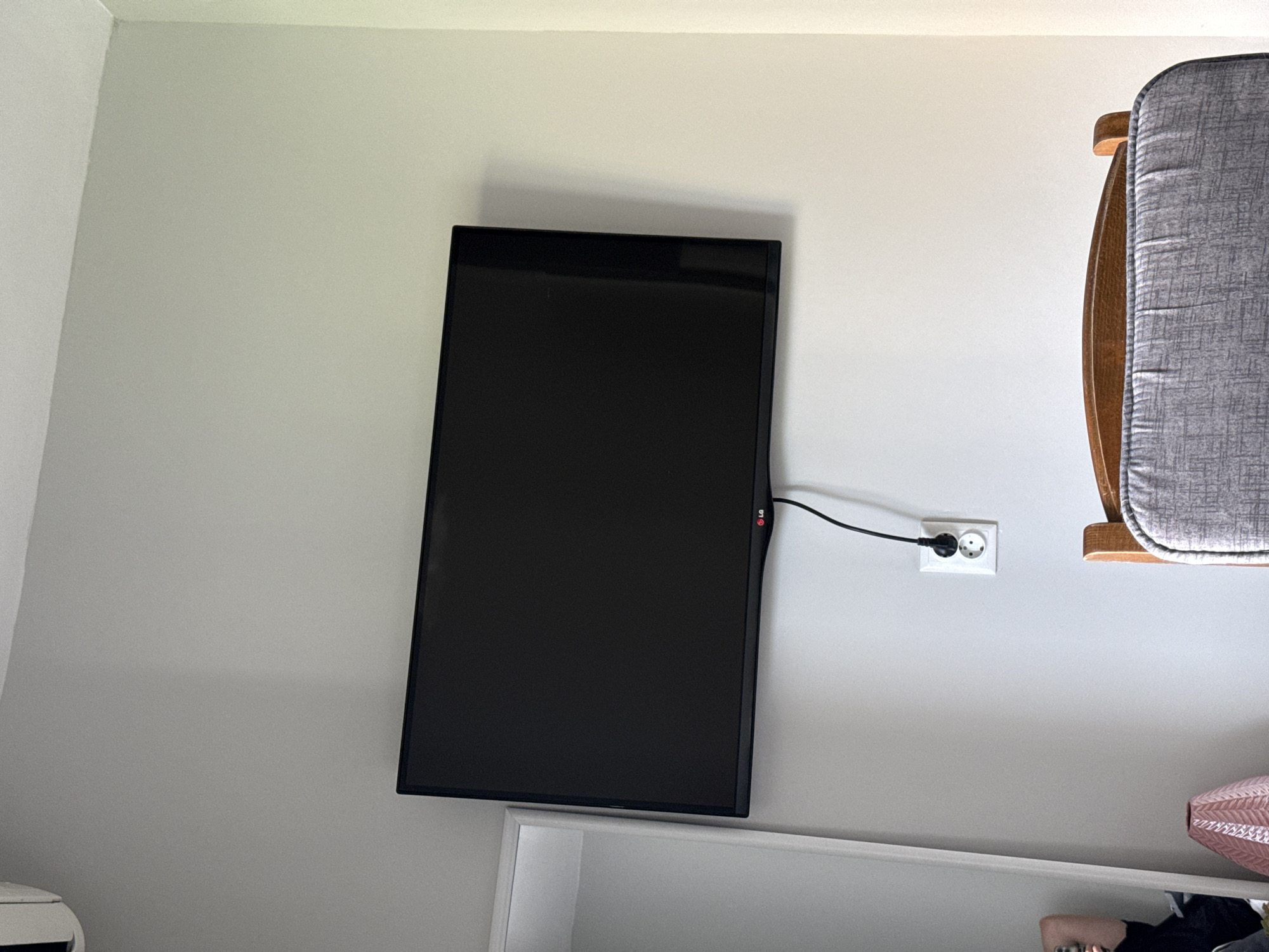 A wall-mounted LG television is plugged into a European-style electrical outlet. The television is mounted above a white shelf or cabinet. Part of a wooden chair with a gray cushion is visible to the right. The background is a plain white wall. The location is not identifiable from this image alone and therefore I cannot confirm it to be in Stara Zagora, Bulgaria.