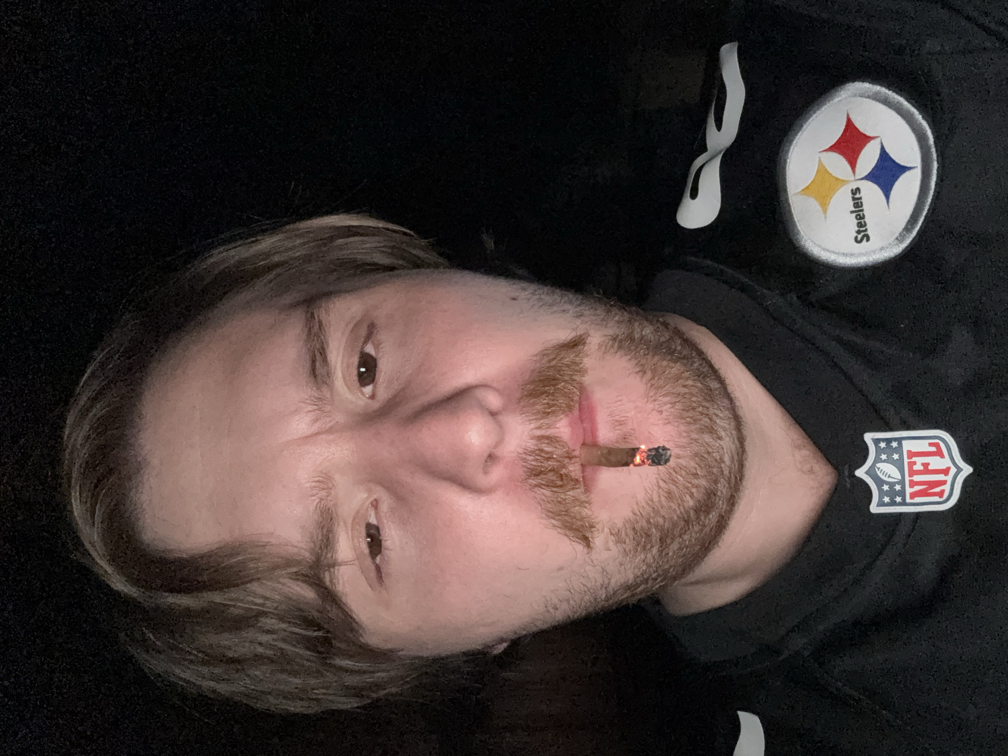 A man wearing a black Pittsburgh Steelers NFL jersey with the team logo on the right chest and an NFL shield logo on the left chest, is pictured from the chest up, against a dark background. The man has light-colored skin, brown hair, and a mustache. He is holding a lit cigar between his lips, with ash visible at the end. The man's eyes are looking towards the camera with a slight frown. The lighting suggests the photo was taken indoors or at night. There is no identifiable indication of the city of Cañada de Madero, Mexico in the image.