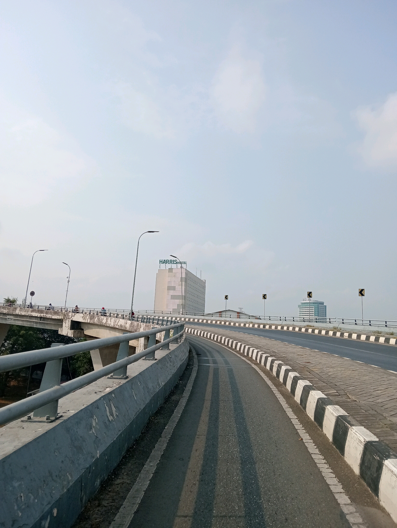This is an eye-level, outdoor, vertical shot of a highway overpass in Samarinda, Indonesia. The overpass has a grey concrete railing on the left side of the frame and a black and white striped curb on the right. The road is asphalt and curves to the right. In the background, there is a tall, light-colored building with "HARRIS HOTEL" written in green on top. There are also two other buildings, one green and one light brown, further in the background. Several streetlights are visible along the overpass. The sky is a pale blue with a few wispy clouds. A few people on motorcycles can be seen on the overpass in the distance.