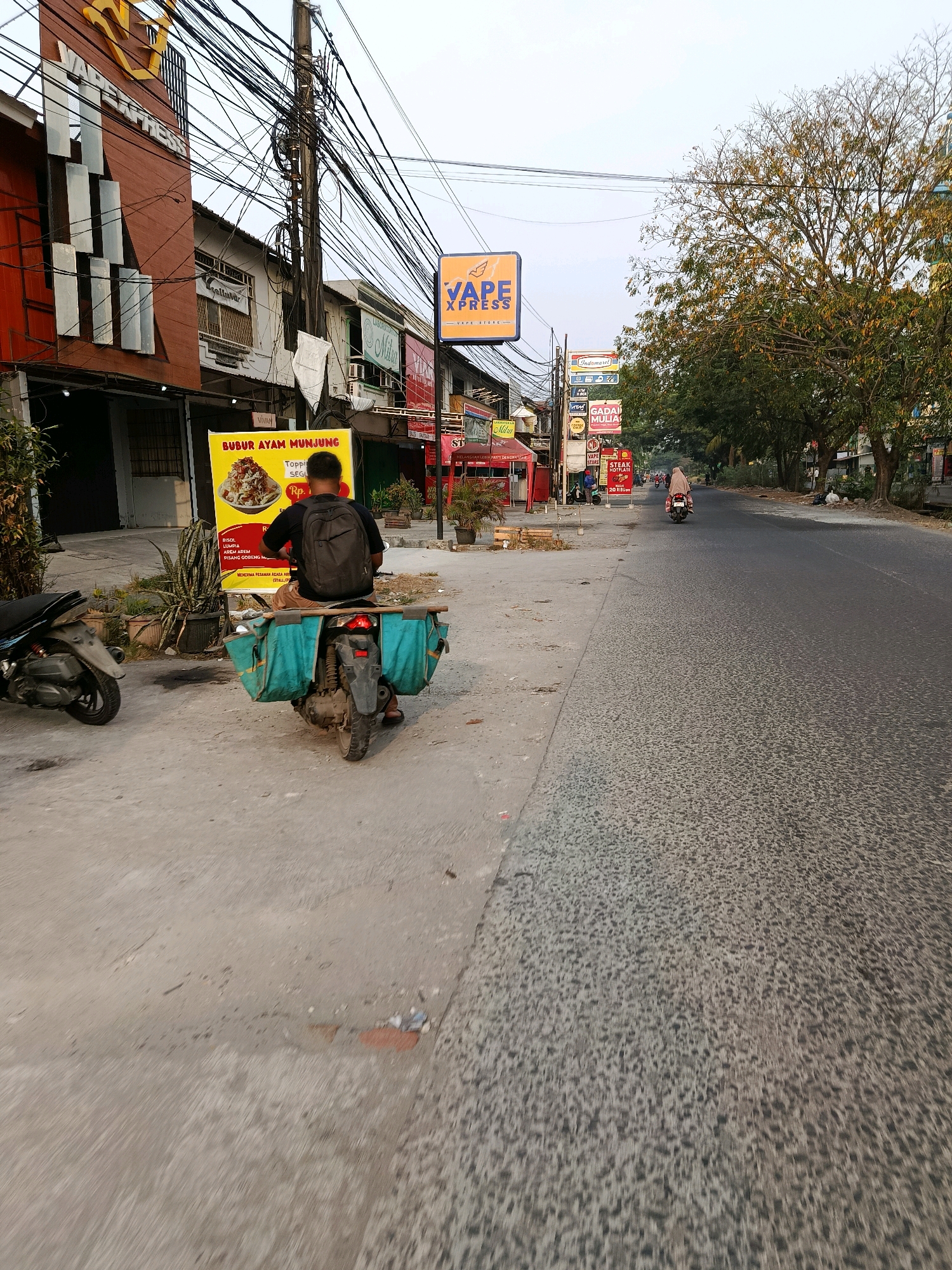 The image depicts a street scene in Bekasi, Indonesia. In the foreground, a man wearing a black t-shirt and brown shorts sits on a motorcycle. The motorcycle has two large, turquoise saddlebags attached to the rear. The man is facing away from the camera, looking towards a yellow banner advertisement. The banner features text in Indonesian, including "BUBUR AYAM MUNJUNG" (Chicken Porridge Munjung) and prices. To the left of the man, another motorcycle is parked. The street itself is paved and appears to be in a commercial or residential area, with buildings visible along the left side of the road. One building has a sign with "VAPE EXPRESS" written on it. Overhead, a complex network of electrical wires is visible. Further down the street, other buildings and trees line the roadside. A person on a motorcycle is seen riding away from the camera in the distance. The sky is overcast.
