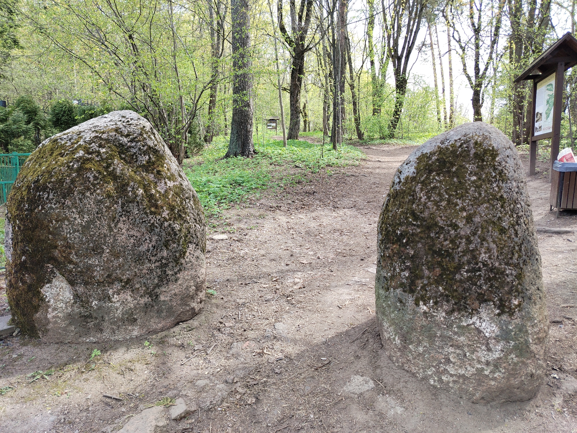 Two large, moss-covered boulders flank a dirt path in a wooded area. The path leads into the distance, with various trees and foliage visible. In the background, there is a wooden information board and a trash receptacle. The setting appears to be a park or natural area within the city of Unknown, Unknown.