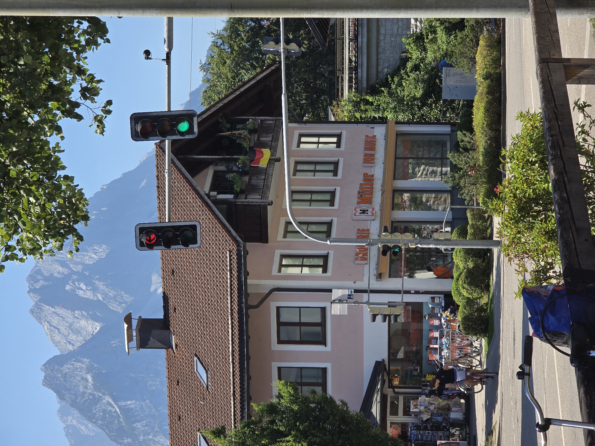 The image captures a street scene in Garmisch-Partenkirchen, Germany, on a clear, sunny day. In the foreground, a wooden fence runs horizontally across the bottom of the frame, with lush green bushes and vegetation beyond it. To the left, the handlebars and front wheel of a bicycle are visible, equipped with a blue bag. A paved road is in the mid-ground, with a woman on a bicycle riding away from the camera on the right side of the road.
Dominating the mid-ground is a pale pink building with a brown tiled roof and dark brown window frames. Above the entrance, a balcony is adorned with potted plants and a German flag. The building features a sign for "Müller FOR MUSIC". To the left of this building, a shopfront is visible with displays of merchandise. Traffic lights are positioned at intervals above the road, with one showing a red light and another displaying a green light.
In the background, a dense line of green trees separates the buildings from the majestic, jagged mountain peaks that rise sharply against a clear blue sky. The mountains suggest the scenic Alpine setting of Garmisch-Partenkirchen.