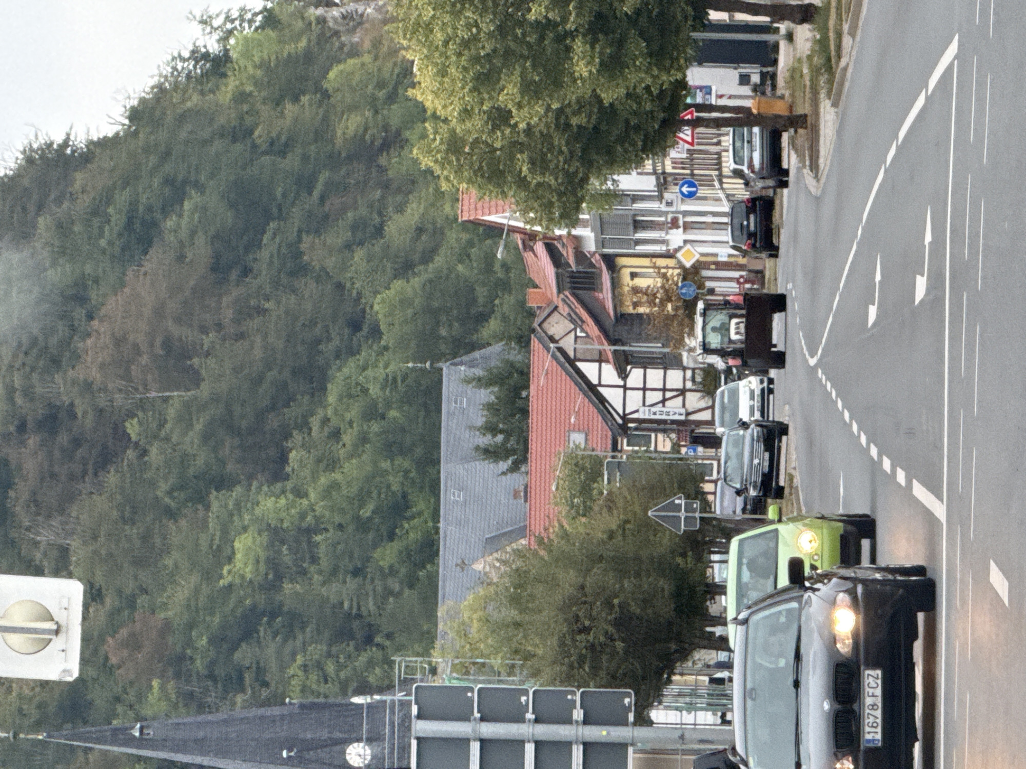 The image depicts a street scene in Niedersachswerfen, Germany. In the foreground, a black BMW sedan with a license plate reading "1678 FCZ" is driving on a road with lane markings. Behind it, a bright green Volkswagen Lupo is also in traffic. Further down the road, a tractor is visible, followed by several other vehicles, including a dark SUV and a white Audi. The street is lined with buildings, some with traditional German architecture, and trees. In the background, a lush, tree-covered hill rises steeply. A church spire is visible on the left side of the image, and a large, modern building with a clock face is mounted above it. The sky is overcast, suggesting a cloudy day. There are several street signs and traffic signals present throughout the scene.