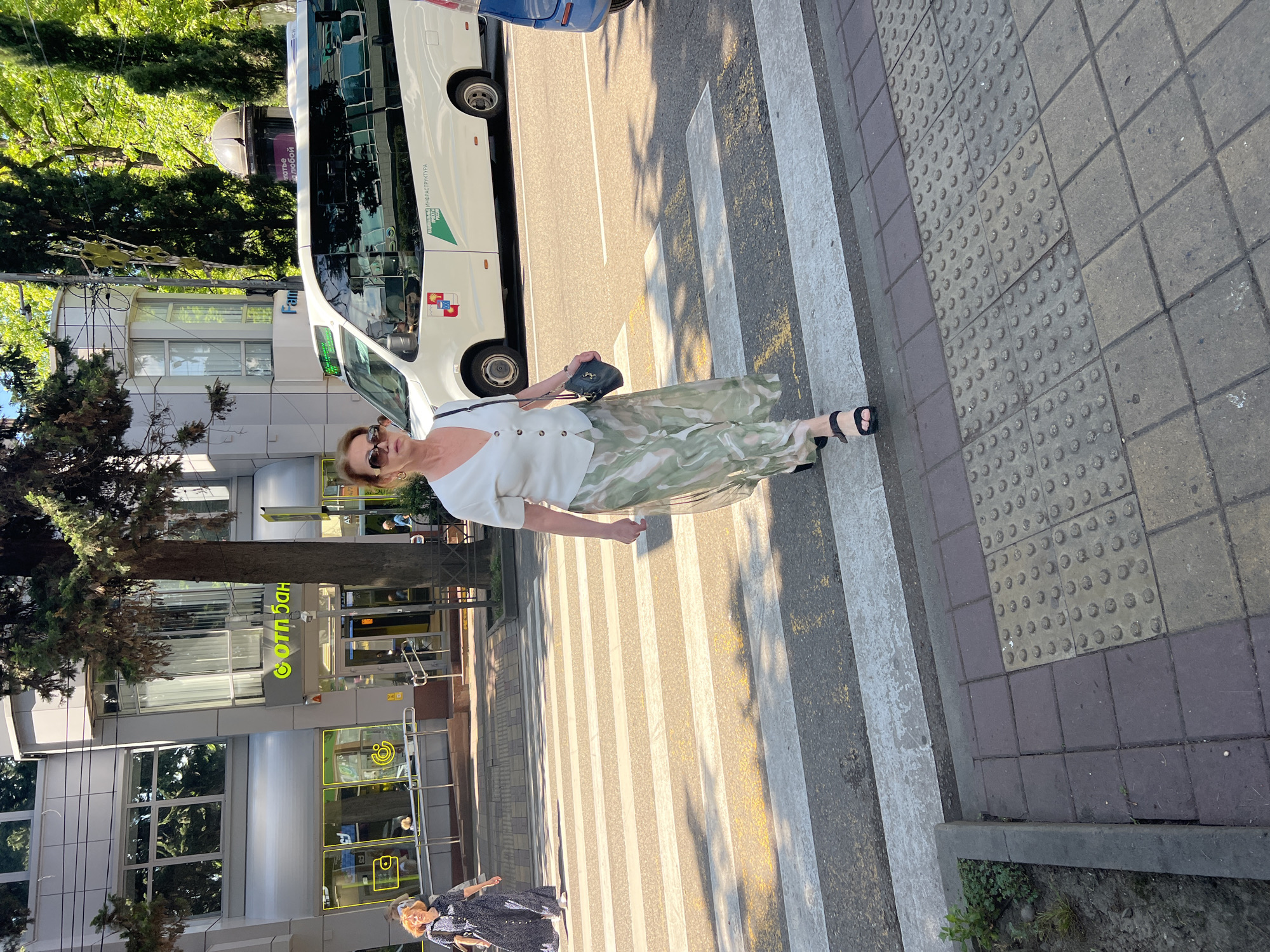 The image captures an urban street scene in Sochi, Russia, on a bright, sunny day, likely late morning or afternoon, as evidenced by the clear sunlight and distinct shadows.
The central subject is a woman with light brown hair, wearing sunglasses, a white short-sleeved top, and a long, flowing skirt with a pattern of green, white, and muted tones. She is wearing black open-toed sandals and carrying a small dark handbag. She appears to be walking on or near a pedestrian crossing, marked by prominent white zebra stripes on the road.
In the background, a large white bus with reflective windows is visible on the street, displaying the Russian text "ИНФРАСТРУКТУРА" (Infrastructure) and "ЖИЗНЬ В" (Life in), alongside a green leaf-like logo and another shield-shaped emblem. A portion of a blue car is also seen behind the bus.
To the left, multi-story modern buildings with large windows line the street. One building features a prominent green sign with the Russian text "СотБанк" (SotBank). Another sign further down the street might read "ШОСОС" or "ШОКОС". Lush green trees and bushes are scattered throughout the scene, suggesting a well-vegetated urban area. The sidewalk in the foreground is paved with gray and brown square tiles, including tactile paving with raised dots for the visually impaired, located adjacent to the pedestrian crossing. A second person, dressed in a dark patterned outfit, is partially visible further down the street on the pedestrian crossing. The scene conveys a casual, everyday moment in a city setting.