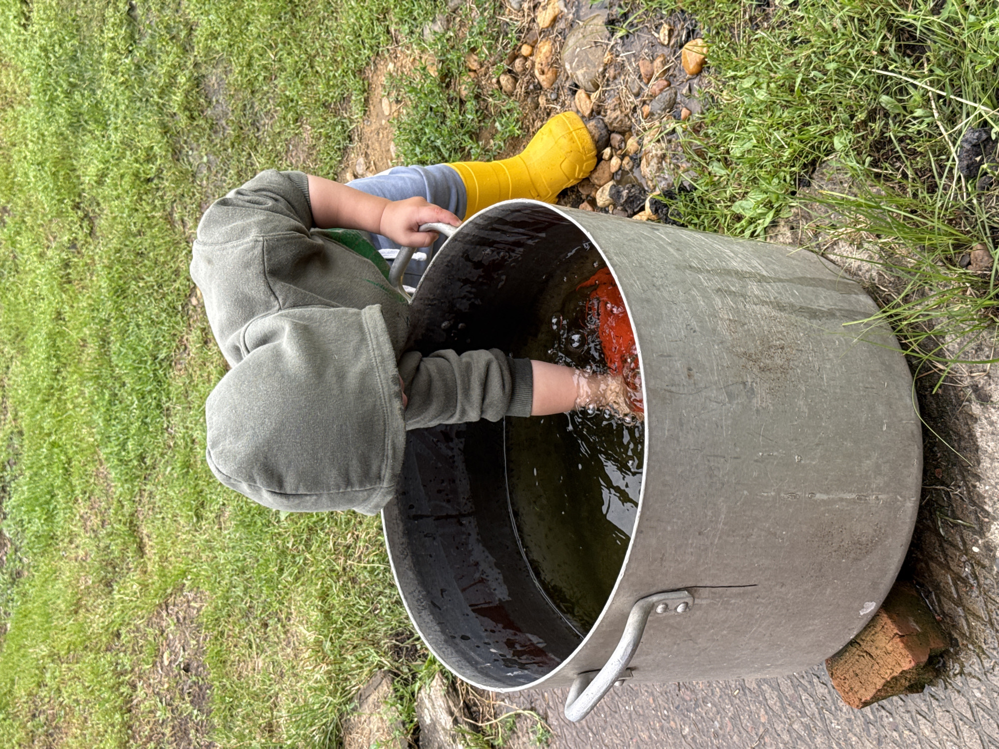 A young child, wearing a green hooded sweatshirt and grey pants, with bright yellow rain boots, is bending over a large, metal pot filled with murky water. The child's arm is submerged in the water, reaching for a red object. The pot is resting on a wet, paved surface, with a single red brick propping up one side. The surrounding environment is grassy with patches of soil and small stones. The scene appears to be outdoors, likely in a backyard or garden area. The location is in Verkhnyaya Uryakh, Russia, given the context.